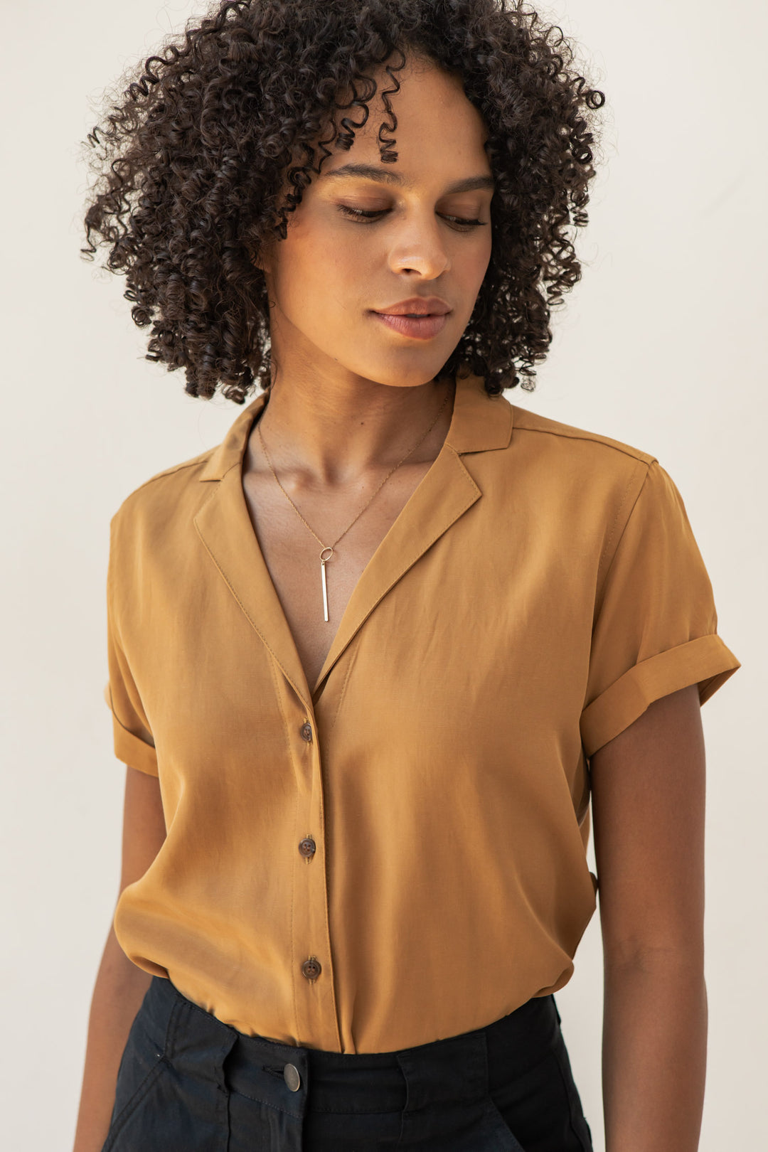 A person with curly hair wears the Bridge & Burn Innes Shirt in Gold—an organic cotton short-sleeve blouse with a relaxed fit—styled with a pendant necklace, looking down neutrally against a plain, light background.