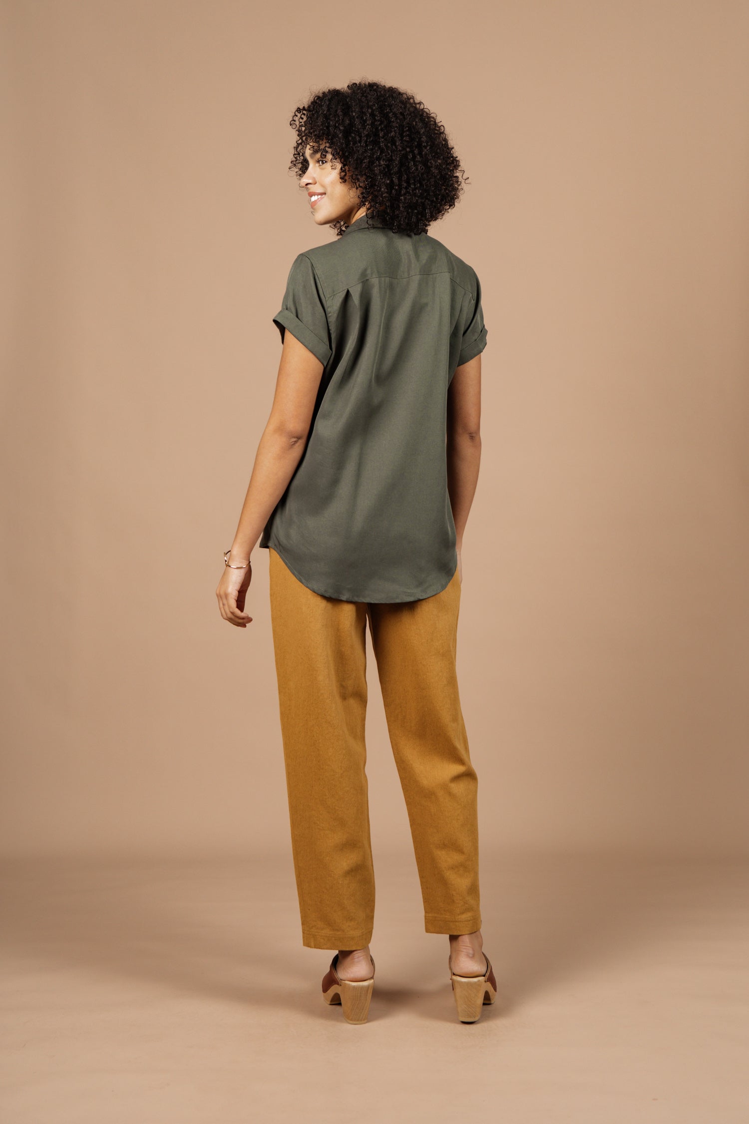 Someone with curly hair, wearing the Bridge & Burn Innes Shirt in Olive, mustard-colored pants, and heeled sandals stands facing away, glancing over their shoulder against a plain beige background.