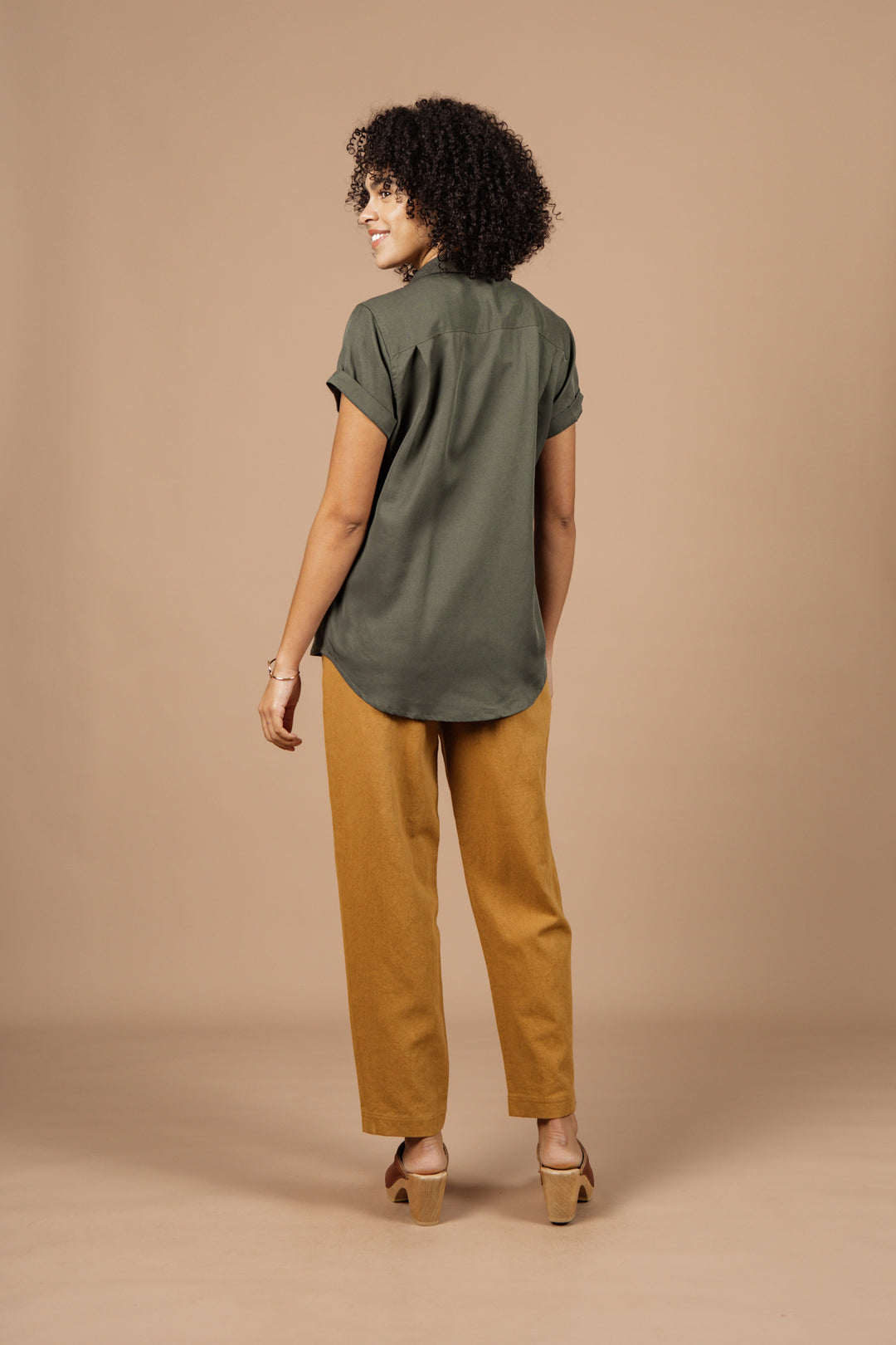 Someone with curly hair, wearing the Bridge & Burn Innes Shirt in Olive, mustard-colored pants, and heeled sandals stands facing away, glancing over their shoulder against a plain beige background.