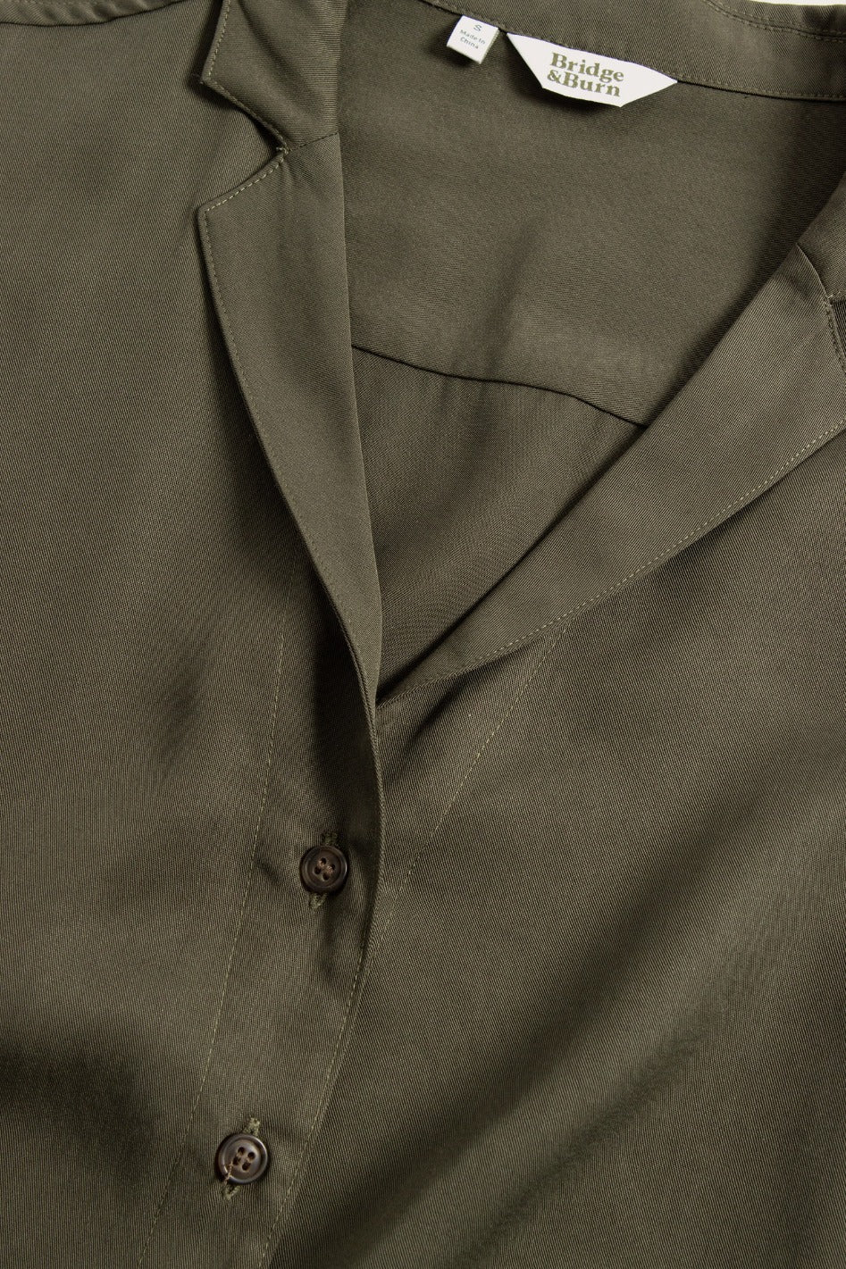 Close-up of the Bridge & Burn Innes Shirt in olive—a relaxed fit button-up with three buttons fastened, a collared neckline, lightweight fabric, and a white tag at the collar showing the brand name.