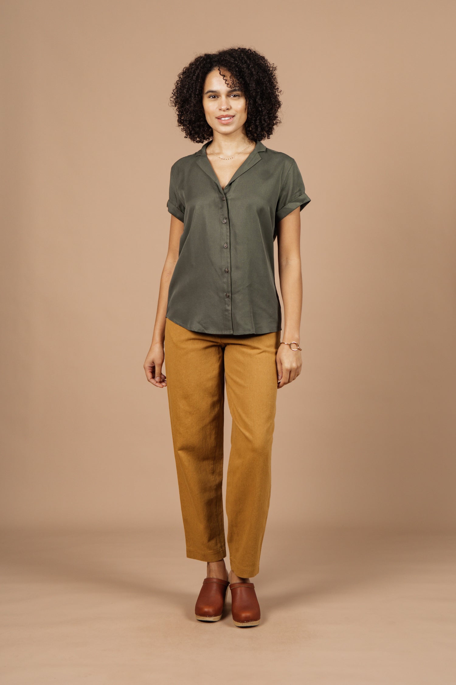 A woman with curly hair and a slight smile stands against a beige background wearing the Bridge & Burn Innes Shirt in olive, mustard yellow pants, brown shoes, and minimal jewelry.