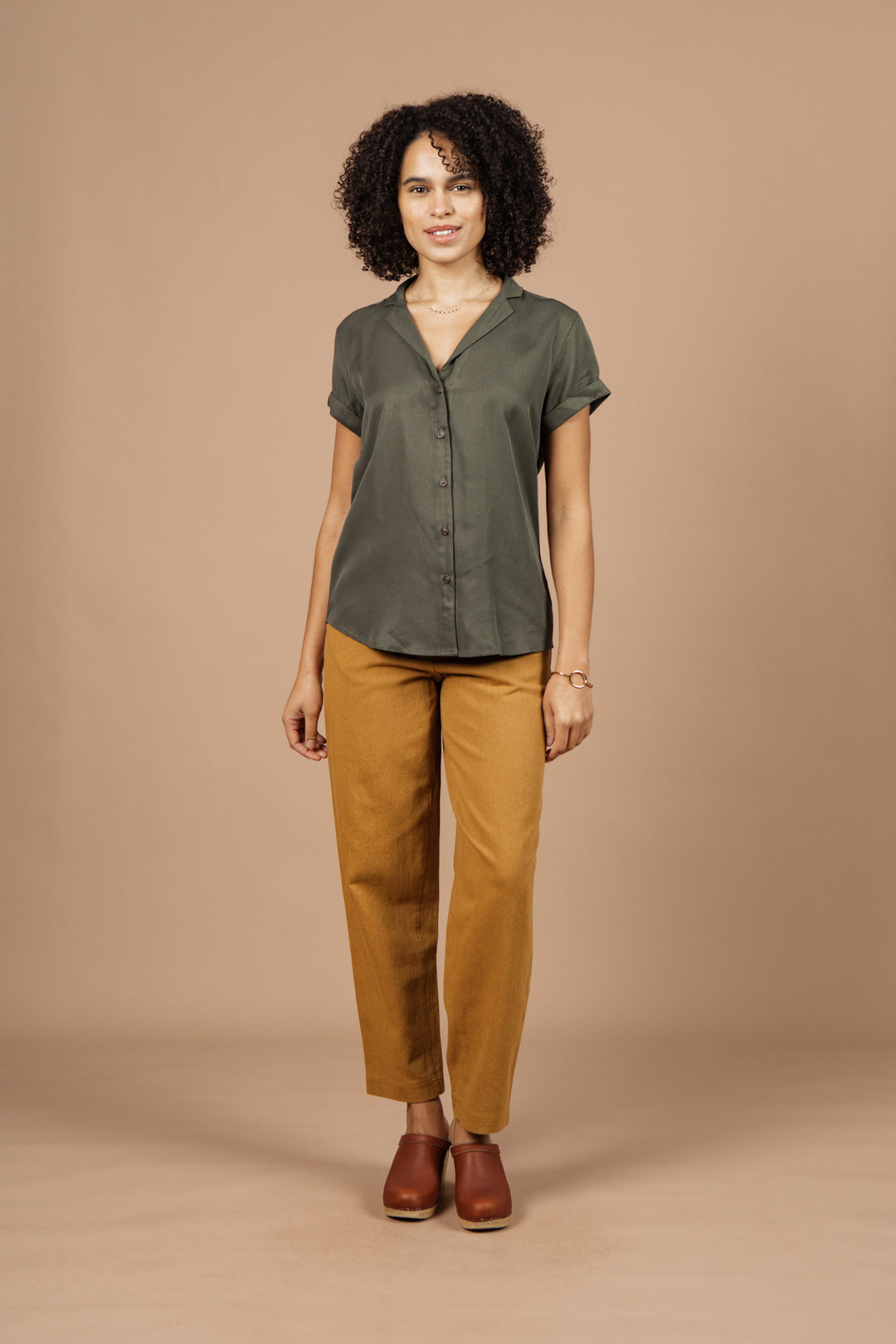 A woman with curly hair and a slight smile stands against a beige background wearing the Bridge & Burn Innes Shirt in olive, mustard yellow pants, brown shoes, and minimal jewelry.