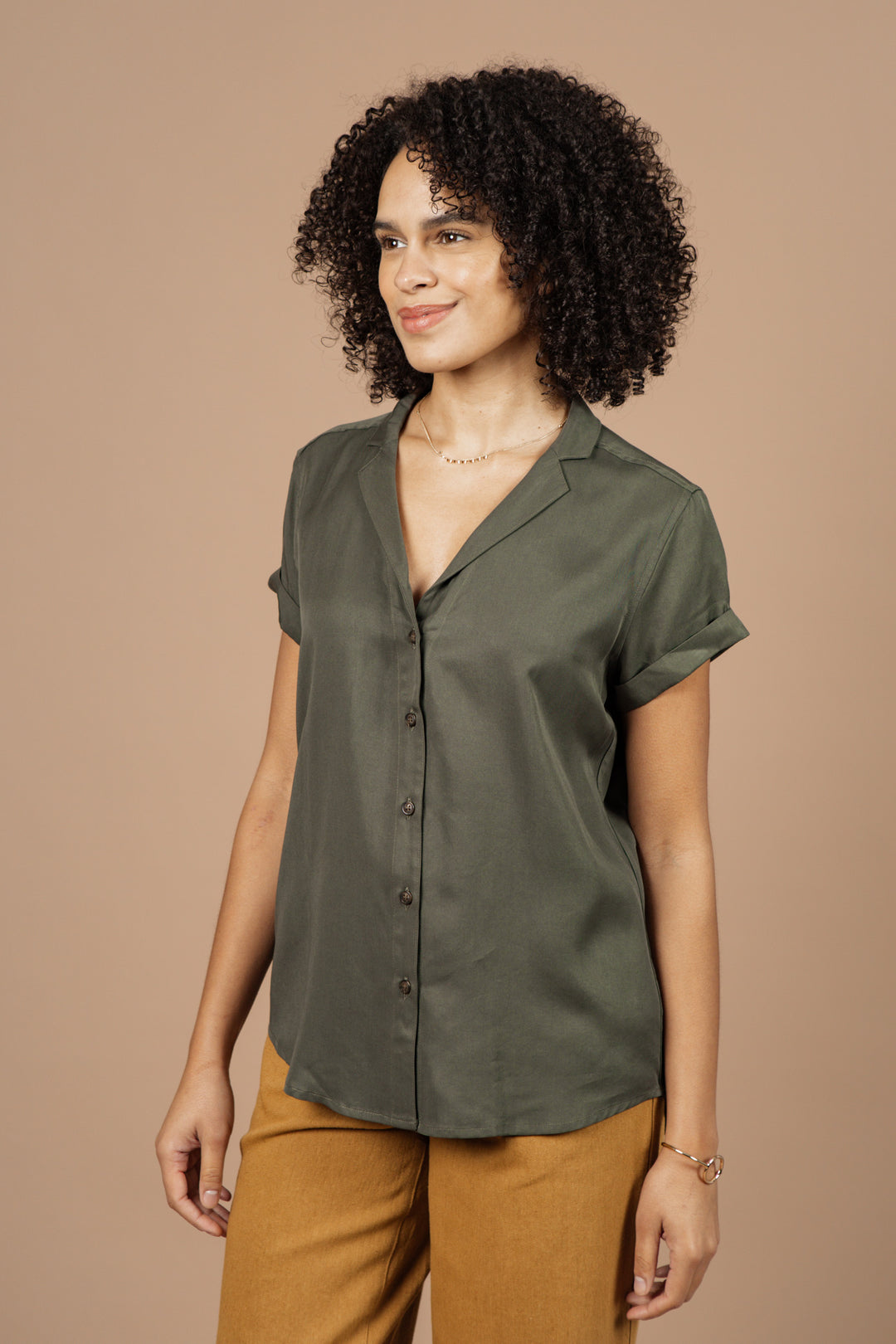 A woman with curly hair smiles gently, wearing the Bridge & Burn Innes Shirt in olive with tan pants, standing against a beige background and looking slightly to the side.