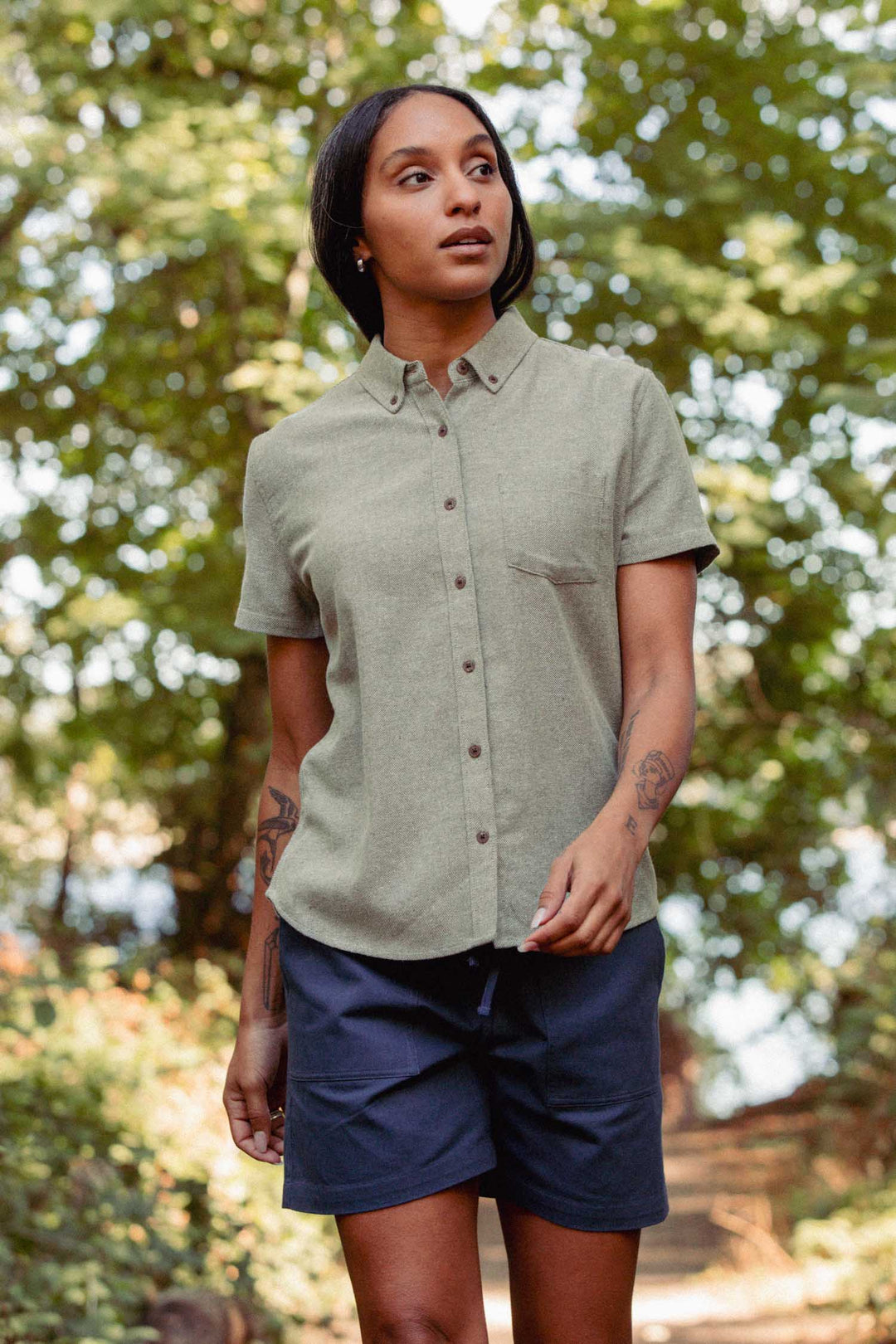 A person with long dark hair stands outdoors on a sunny day, surrounded by lush green trees, wearing the Bridge & Burn Juniper Shirt in Olive Twill paired with dark shorts.