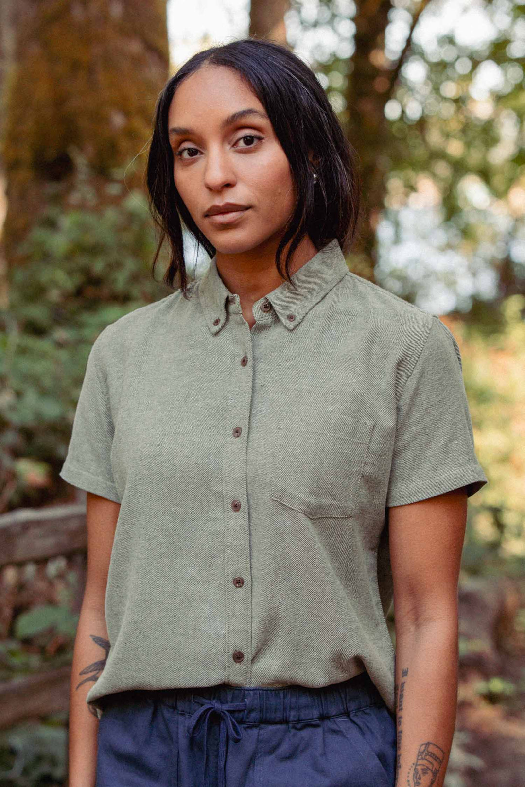 A person with long dark hair stands in a forest wearing the Bridge & Burn Juniper Shirt in Olive Twill and navy pants, surrounded by blurred trees and foliage.