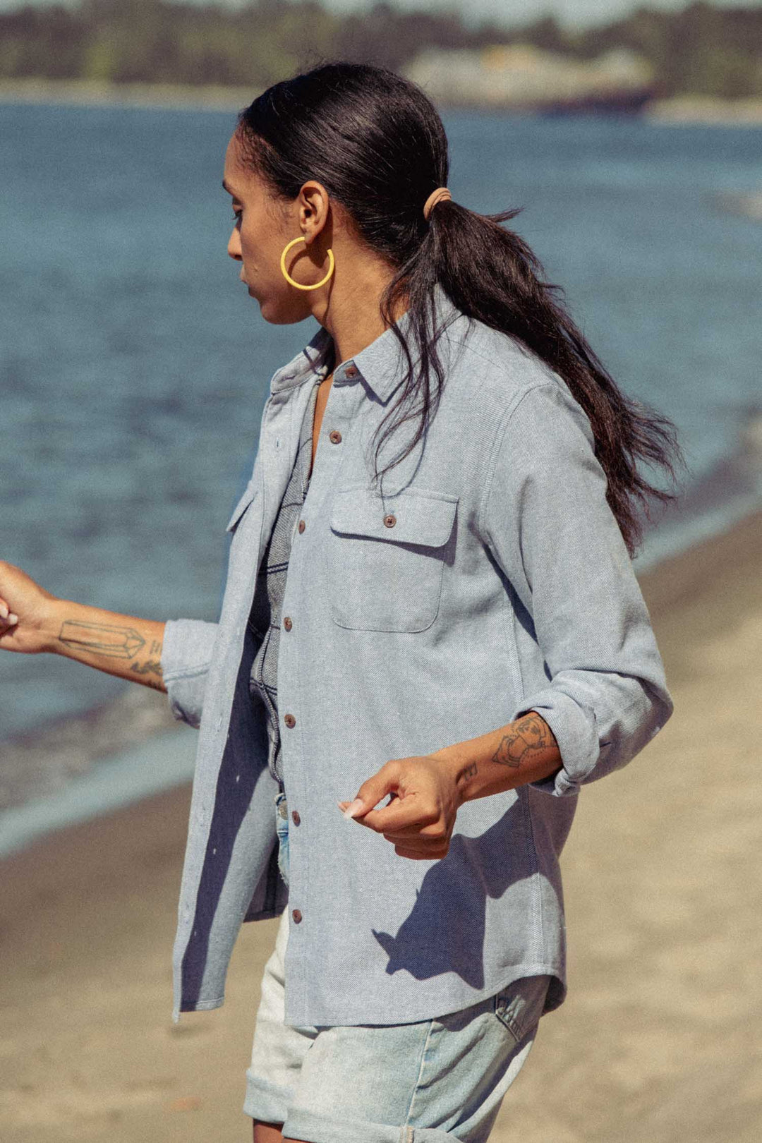 Wearing the Bridge & Burn Lenon Shirt in Denim Twill and shorts, a long-haired person with tattoos and gold hoop earrings strolls along a sandy beach near calm water.