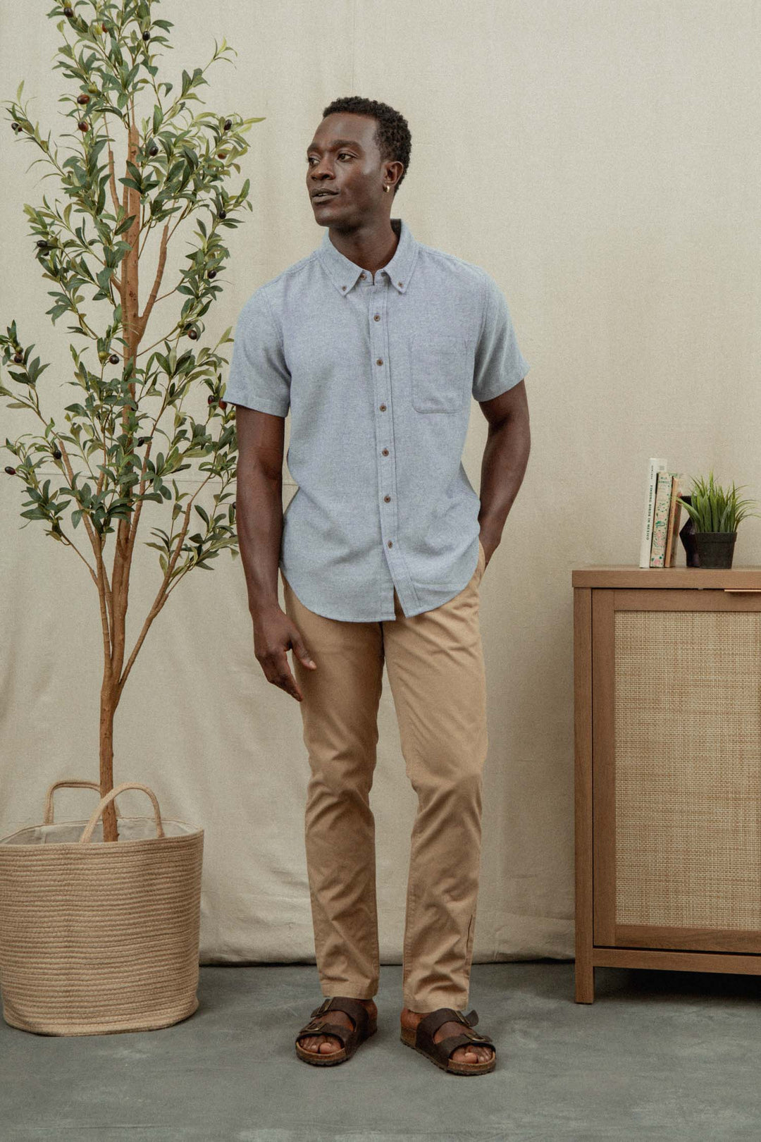 A man stands indoors wearing the Bridge & Burn Bryce Shirt in Denim Twill, paired with tan pants and brown sandals. He is beside a potted plant and a wooden cabinet topped with books and a small plant.