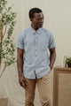 A man wearing the Bridge & Burn Bryce Shirt in Denim Twill and tan pants stands indoors near a plant and cabinet, looking down with one hand in his pocket.