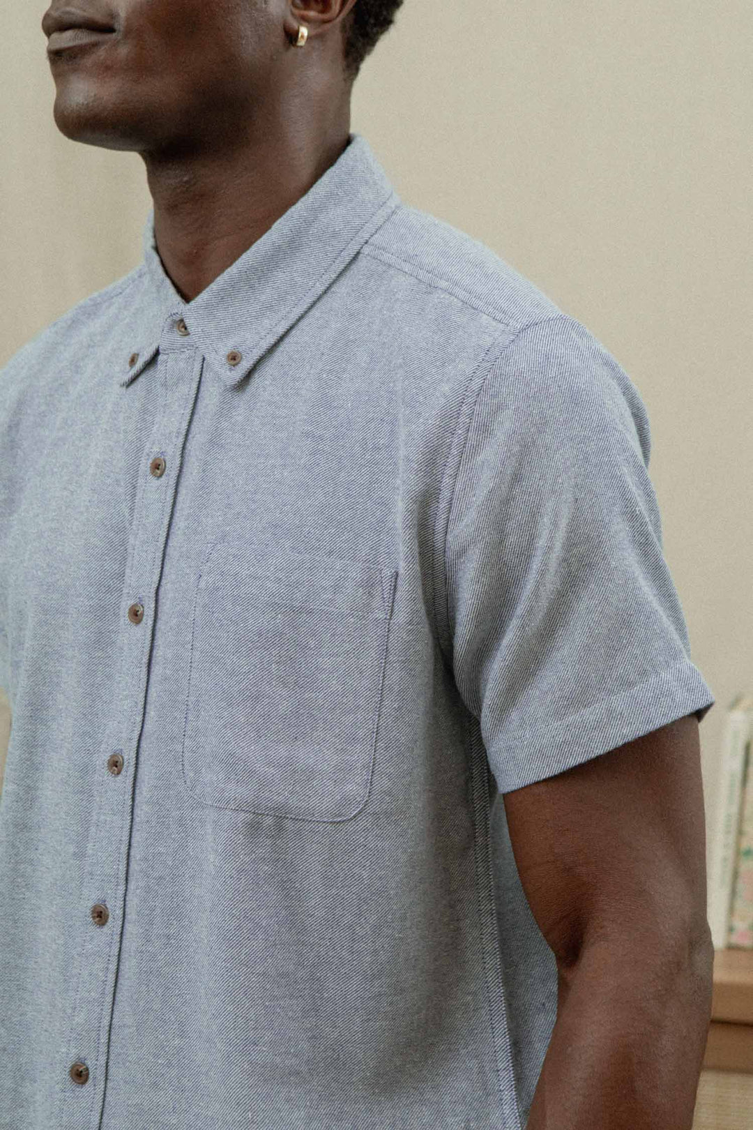 A person wearing the Bridge & Burn Bryce Shirt in denim twill—a light blue short sleeve button-down with a chest pocket—stands indoors, showing only their upper body and part of their face.