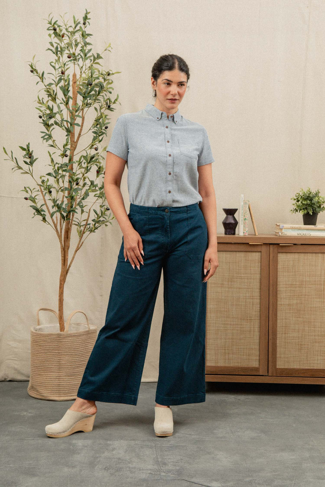 A woman stands indoors wearing the Bridge & Burn Juniper Shirt in Denim Twill, paired with wide-leg dark blue pants and white clogs. She is on a concrete floor, with a plant and a wooden cabinet behind her.