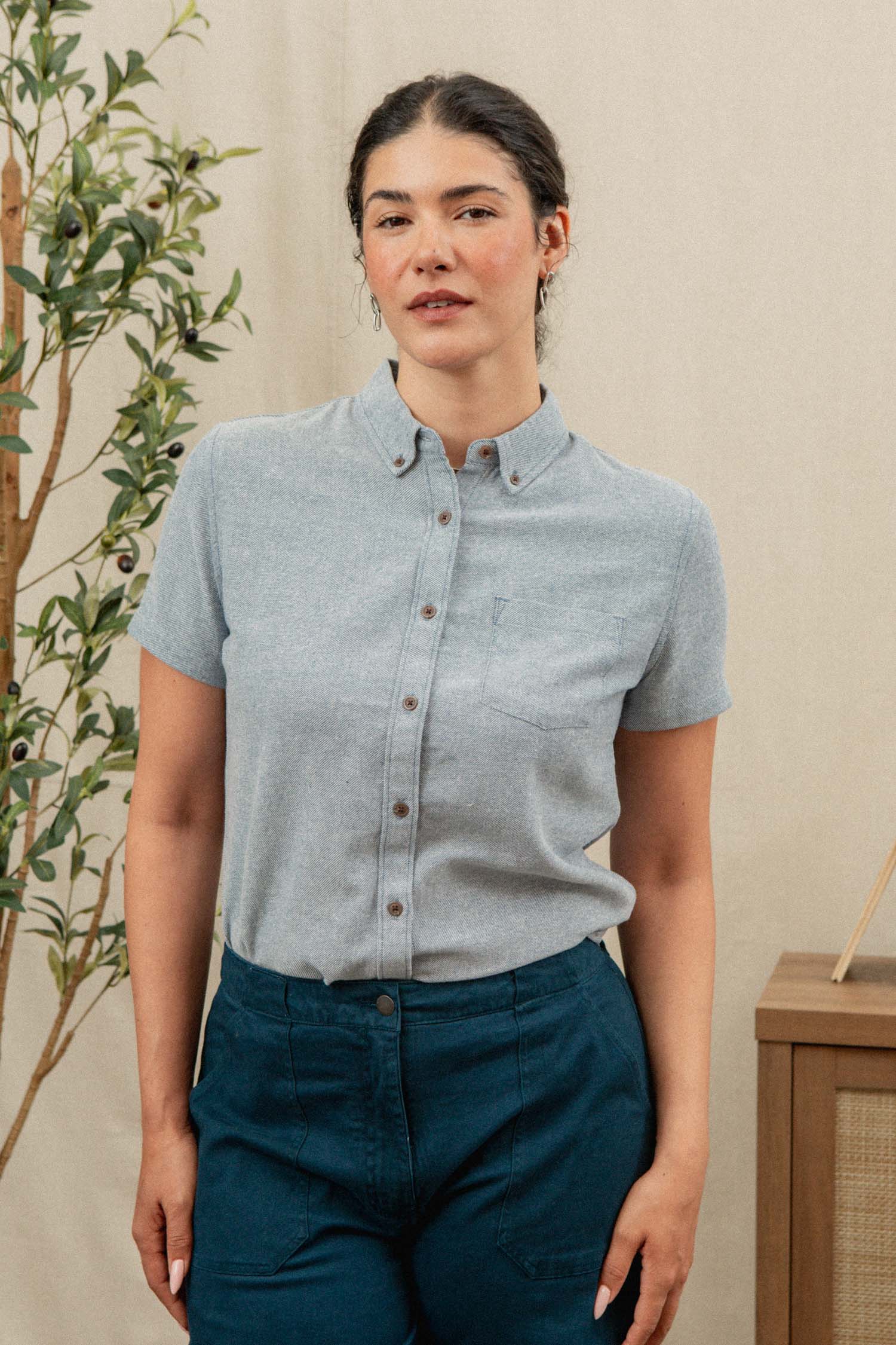 A woman with dark hair pulled back stands indoors, wearing the Bridge & Burn Juniper Shirt in Denim Twill and dark blue pants. A plant and a wooden cabinet are visible in the background.