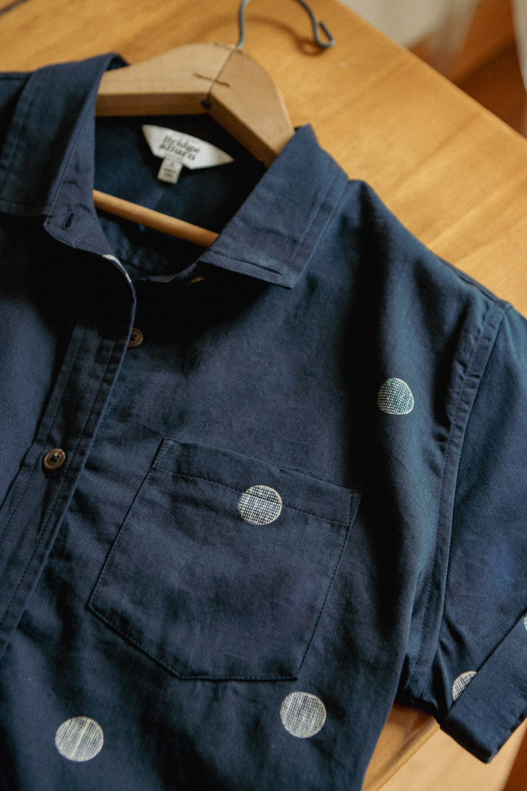 The Bea Shirt / Lunar Polka by Bridge & Burn is a navy blue, short-sleeve, tailored button-down with a front pocket and white polka dots, shown on a wooden hanger over a wooden surface.