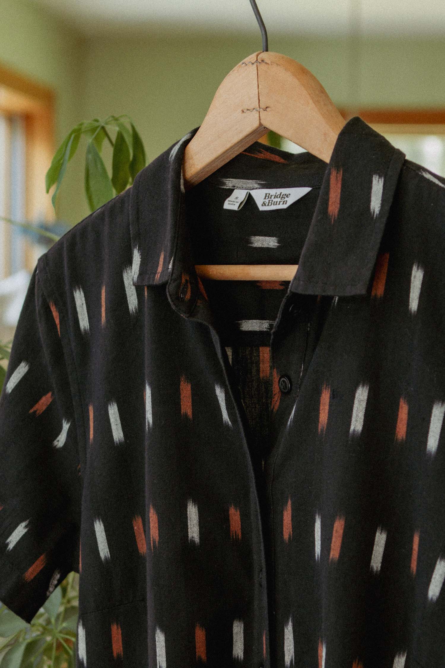 The Delia Dress in Dotted Horizon by Bridge & Burn, featuring black fabric with white and reddish vertical brushstroke-like patterns, hangs on a wooden hanger indoors amid green plants and sunlit windows.
