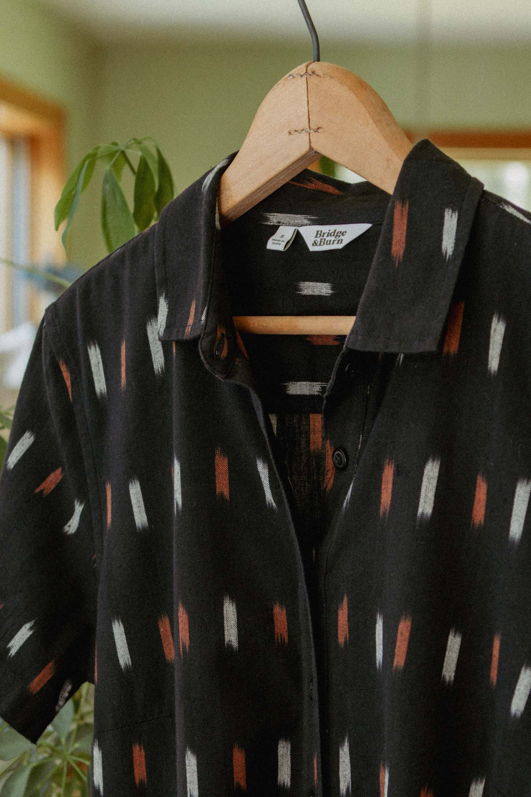 The Delia Dress in Dotted Horizon by Bridge & Burn, featuring black fabric with white and reddish vertical brushstroke-like patterns, hangs on a wooden hanger indoors amid green plants and sunlit windows.