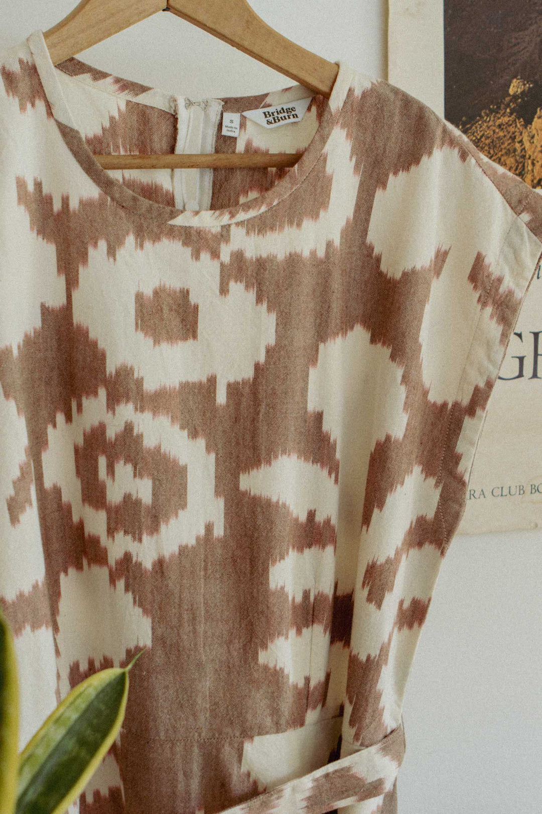 The Eloise Dress / Pop Leaf by Bridge & Burn features a beige and brown abstract geometric pattern. Hanging on a wooden hanger before a white wall and vintage poster, it’s an effortless wardrobe staple for any season.