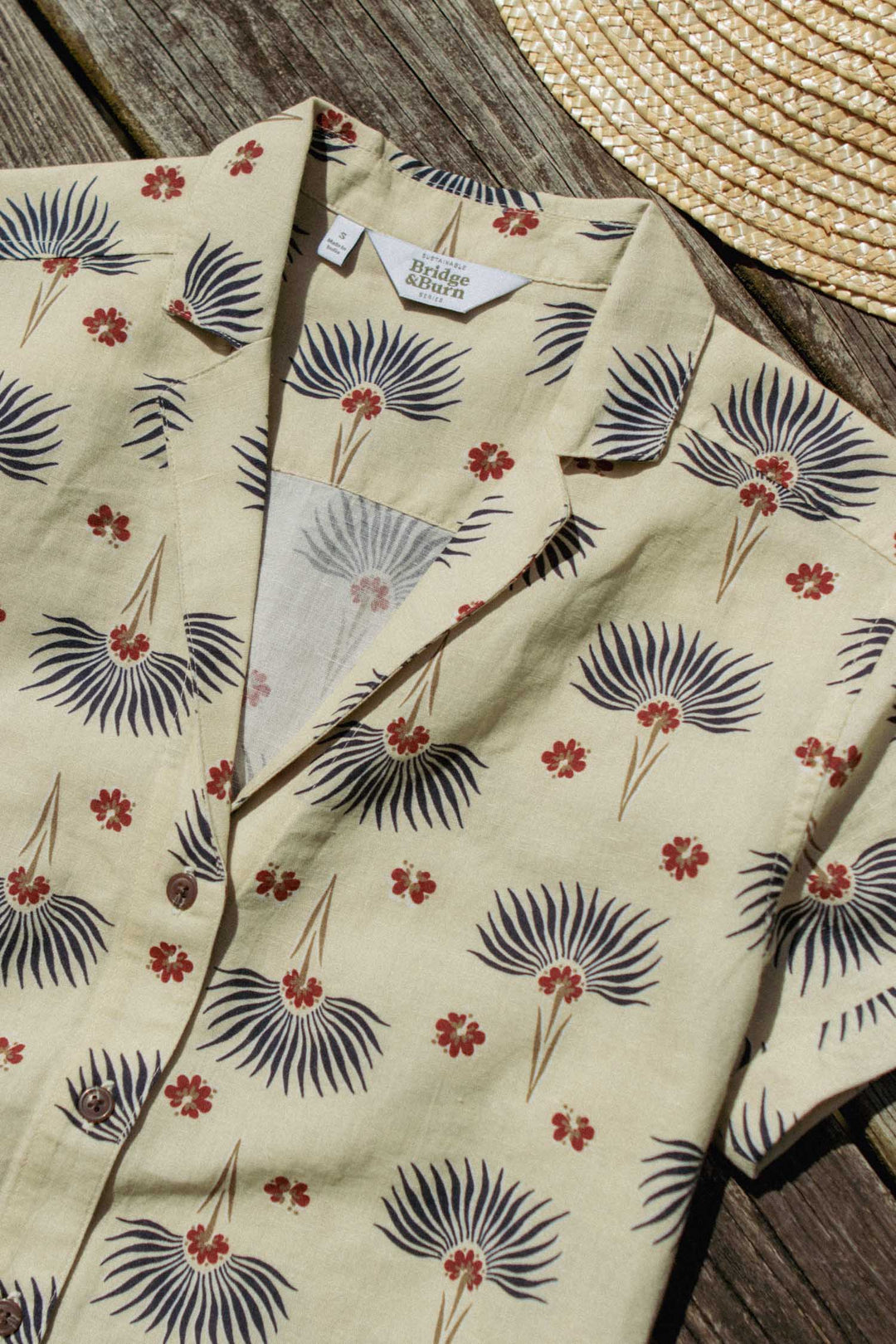 The Bridge & Burn Innes Shirt in Aloha Floral—a beige relaxed-fit linen cotton button-up with a palm leaf and red flower pattern—lies on a wooden surface next to a woven straw hat.