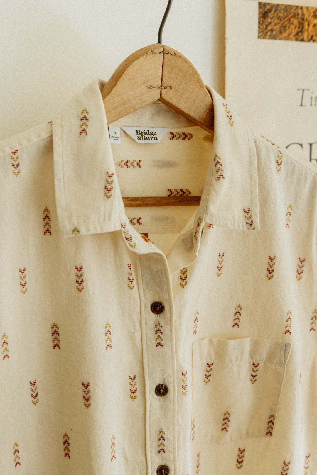 The Bridge & Burn Lana Shirt in Dune Chevron is a cream short sleeve button up with a subtle red and yellow embroidered pattern, made from 100% cotton for comfort. Shown hanging on a wooden hanger against a light wall and partial poster.