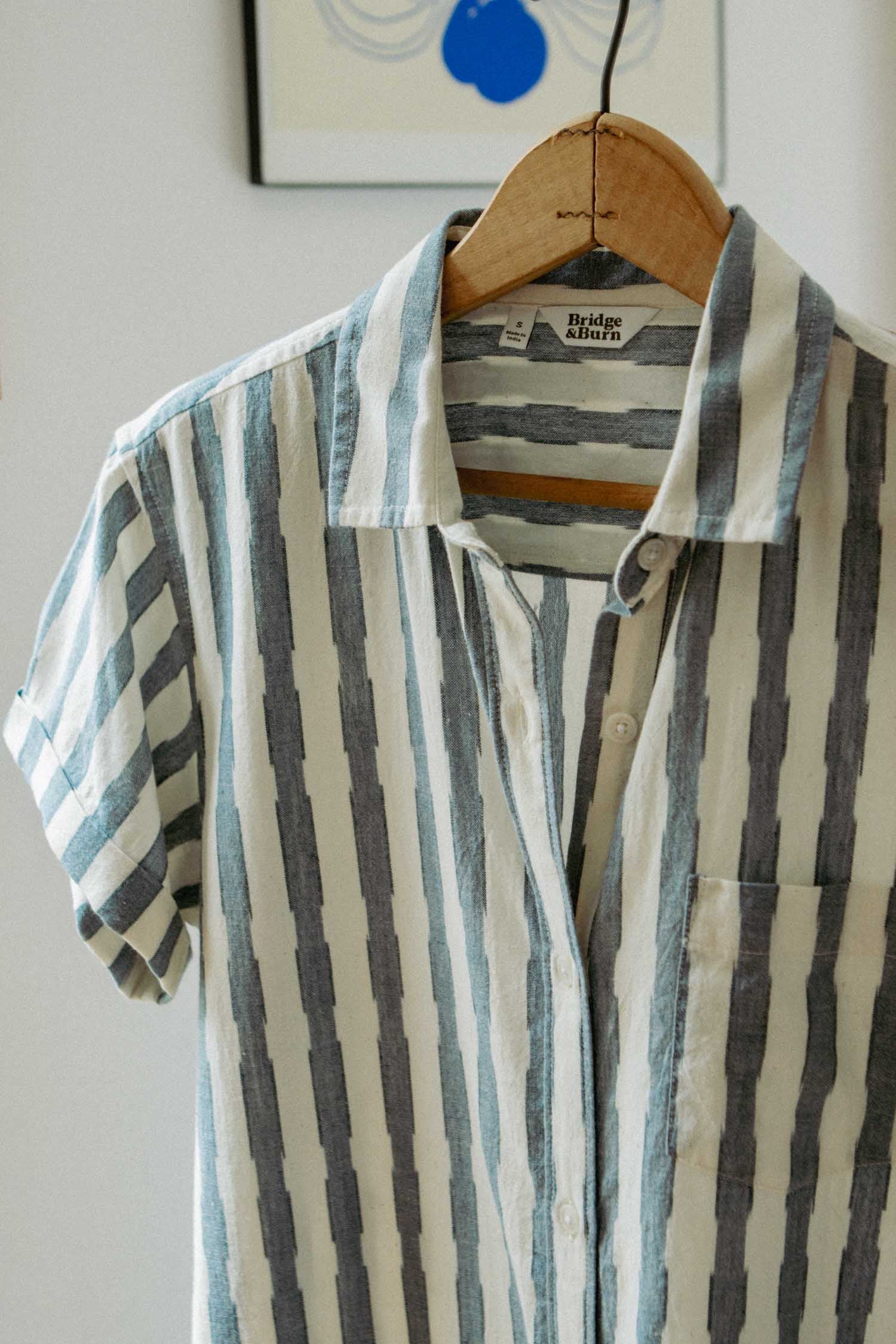 The Bridge & Burn Loren Shirt Dress in Beach Stripe, made from 100% cotton with vertical blue and gray stripes, hangs on a wooden hanger. It features a single chest pocket and a branded label inside the collar.