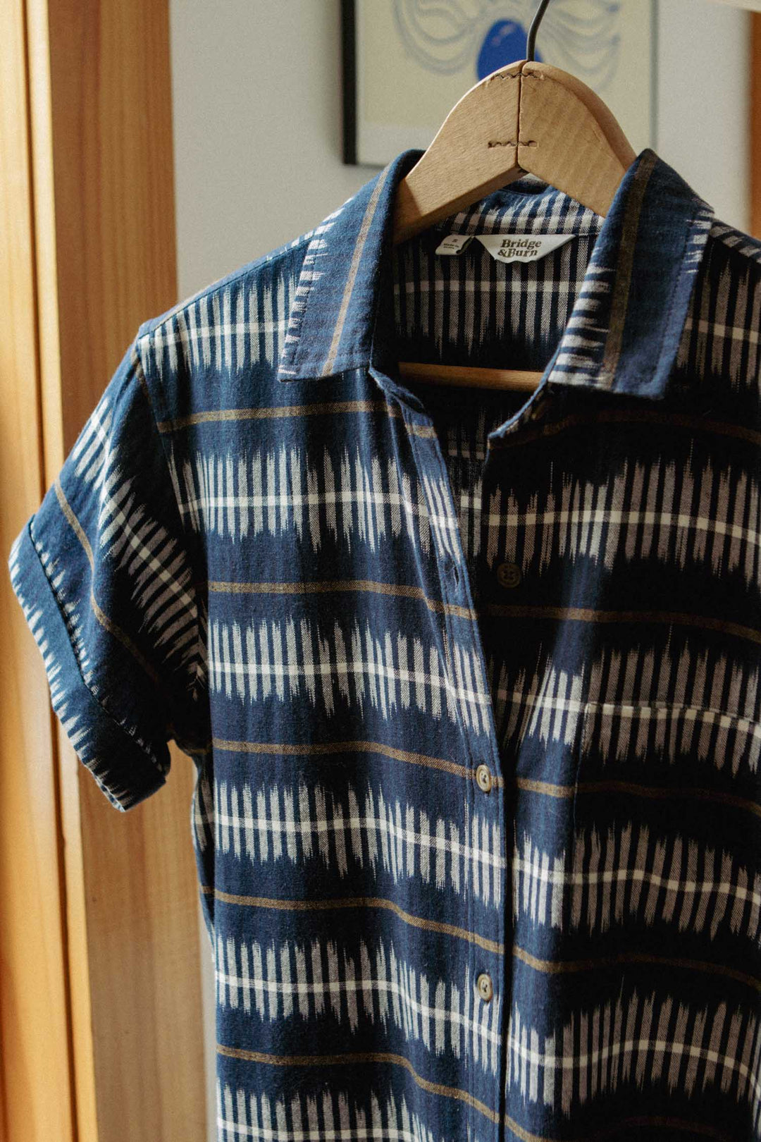 The Loren Shirt Dress in Dusk Mirage by Bridge & Burn, featuring a blue and white geometric pattern and made from 100% cotton, hangs on a wooden hanger in a warmly lit room.