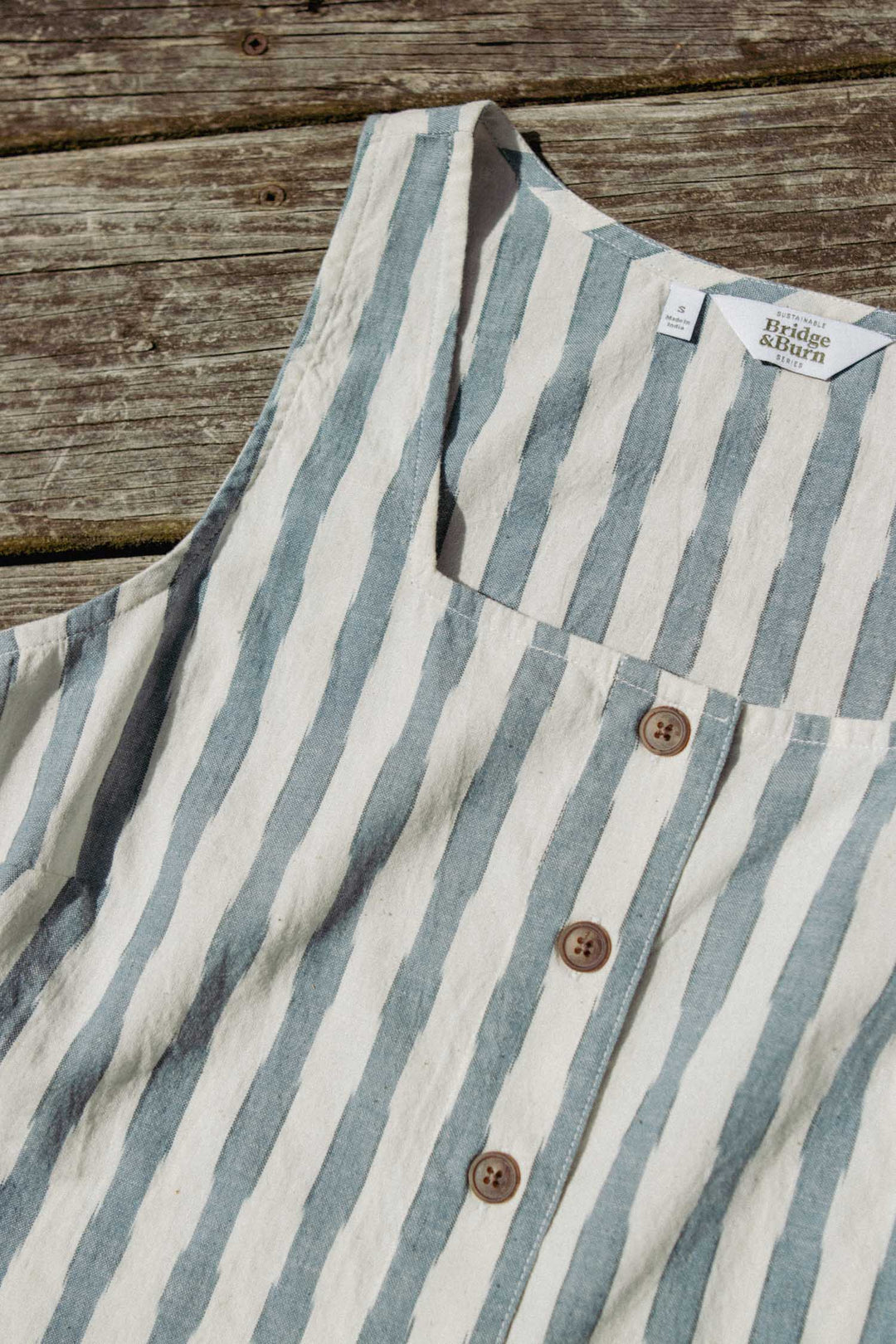 The Sadie Tank in Beach Stripe by Bridge & Burn features vertical blue and white stripes, brown buttons, and a white label, shown laid flat on a wooden surface.