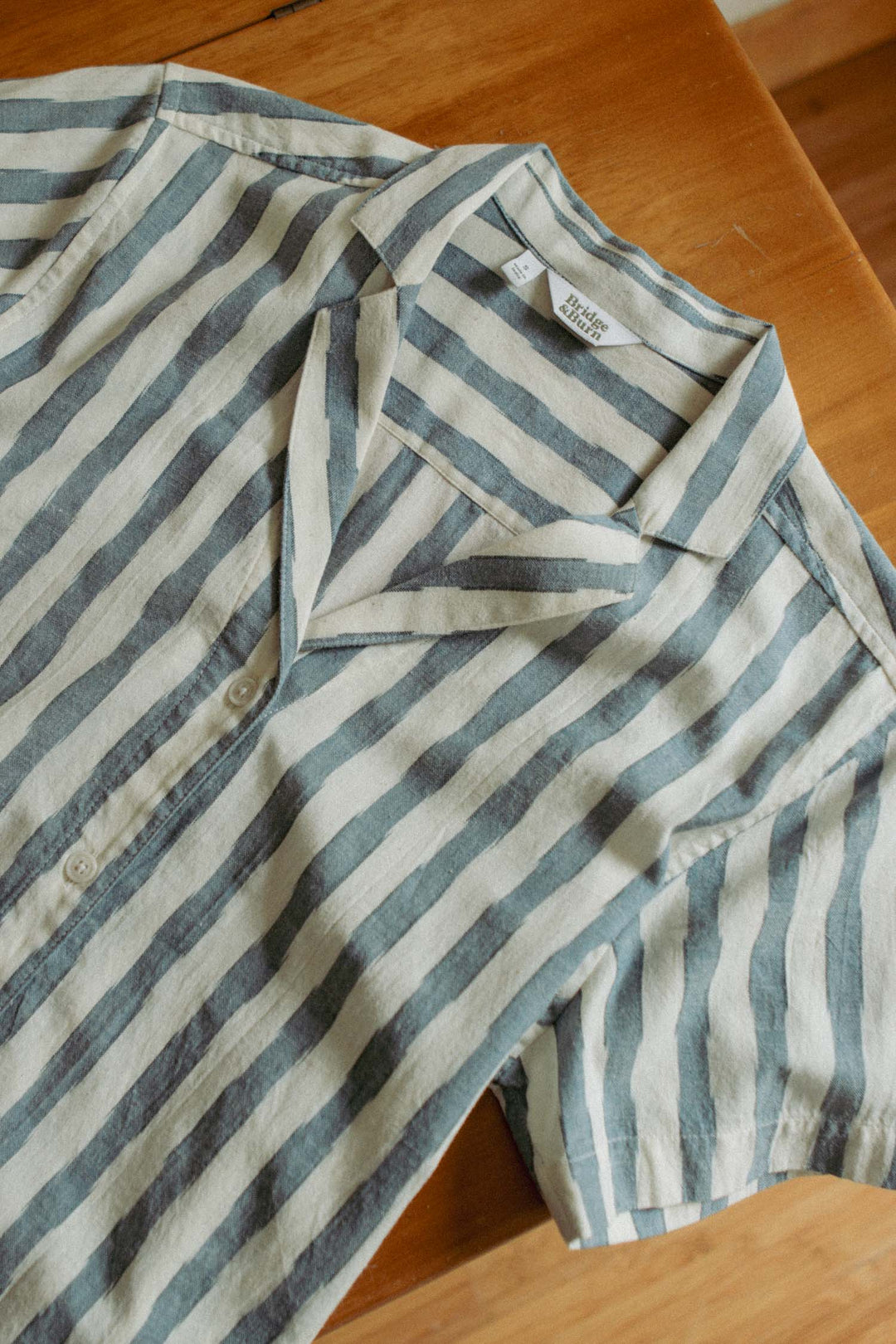 The Wren Shirt / Beach Stripe by Bridge & Burn is a short-sleeved, button-up shirt with a relaxed collar and wide muted blue and off-white stripes. Made from breathable cotton, it's pictured laid flat on a wooden surface.