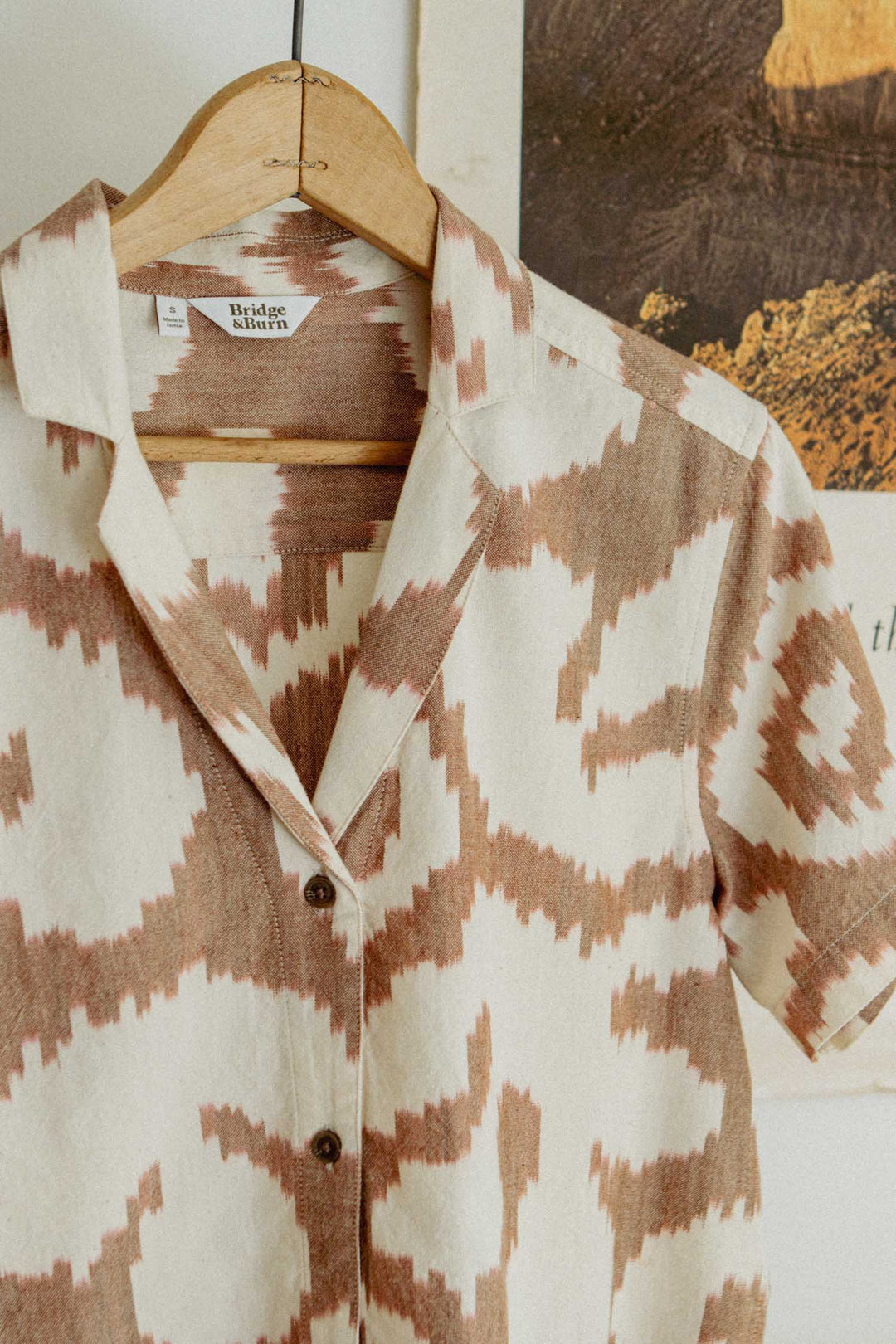 The Bridge & Burn Wren Shirt / Pop Leaf is a short-sleeved, retro-inspired top in beige and brown, perfect for summer comfort thanks to its breathable cotton. Shown on a wooden hanger in front of a partially visible poster or painting.