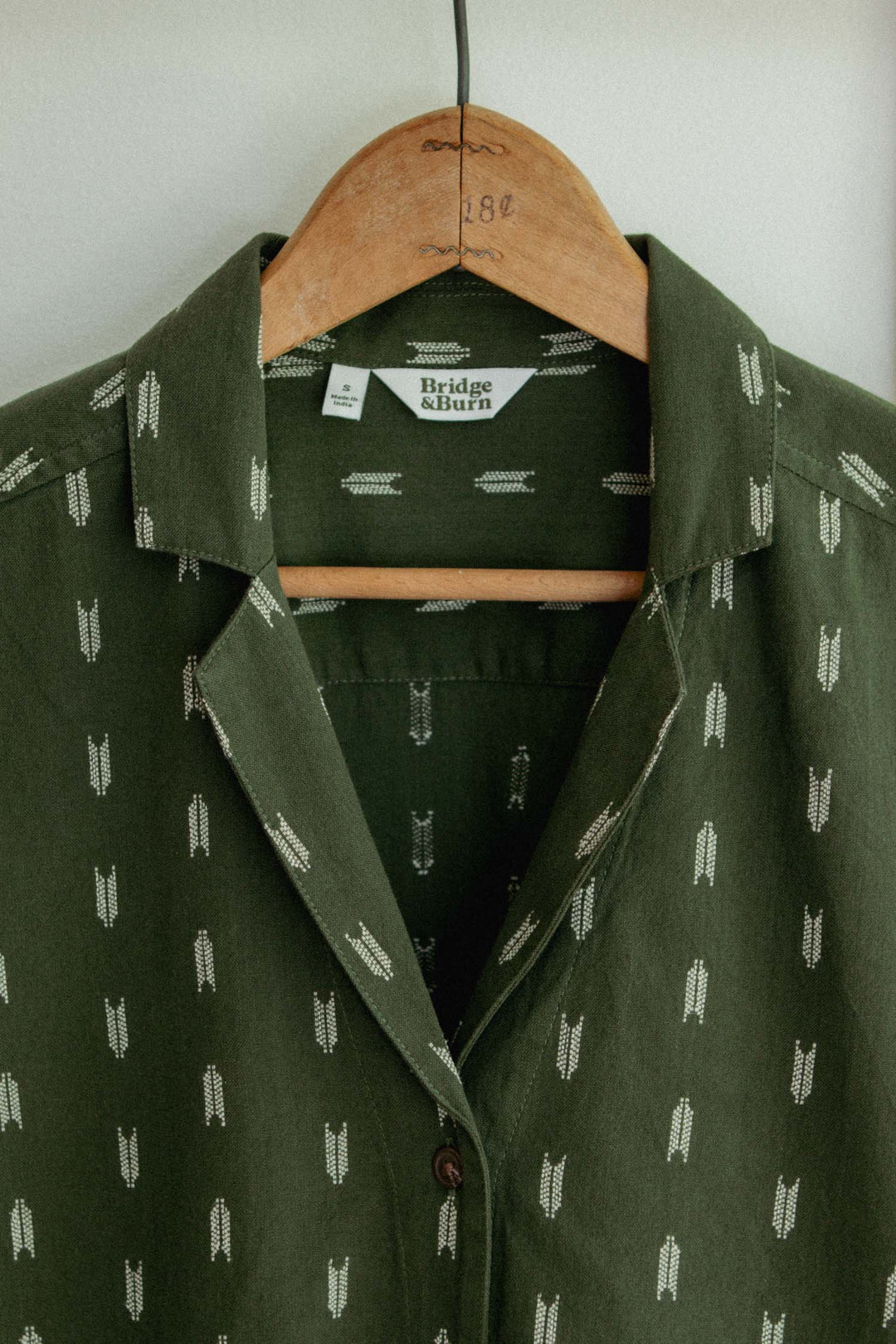 The Bridge & Burn Wren Shirt in Thyme Arrows—a green, relaxed fit button-up with a subtle white arrow pattern—hangs on a wooden hanger. Made from lightweight cotton for summer comfort.