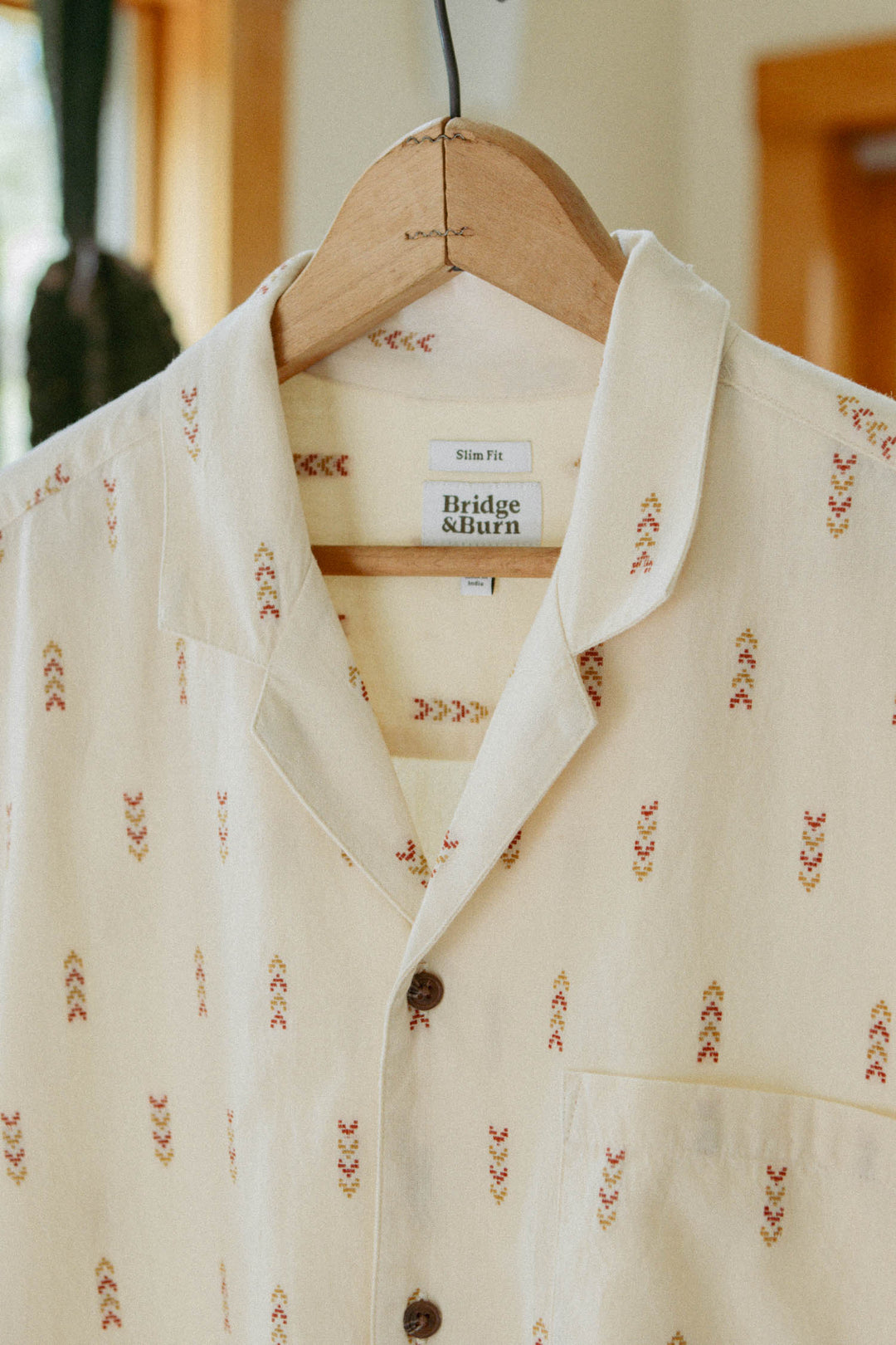 The Collins Slim Shirt in Dune Chevron by Bridge & Burn, a summerweight cotton short-sleeve with a cream camp collar and subtle red and yellow chevron pattern, hangs on a wooden hanger. Label: Slim Fit, Bridge & Burn.
