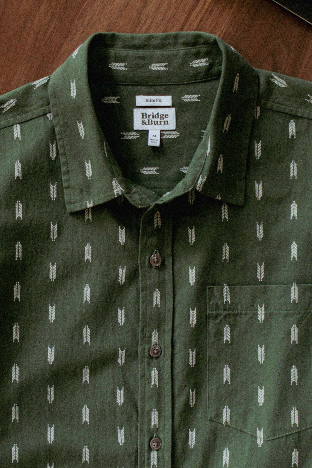 The Bridge & Burn Harbor Slim Shirt in Thyme Arrows features green summerweight cotton with a white arrow pattern, tailored fit, front pocket, and branded collar label, shown laid flat on a wooden surface.