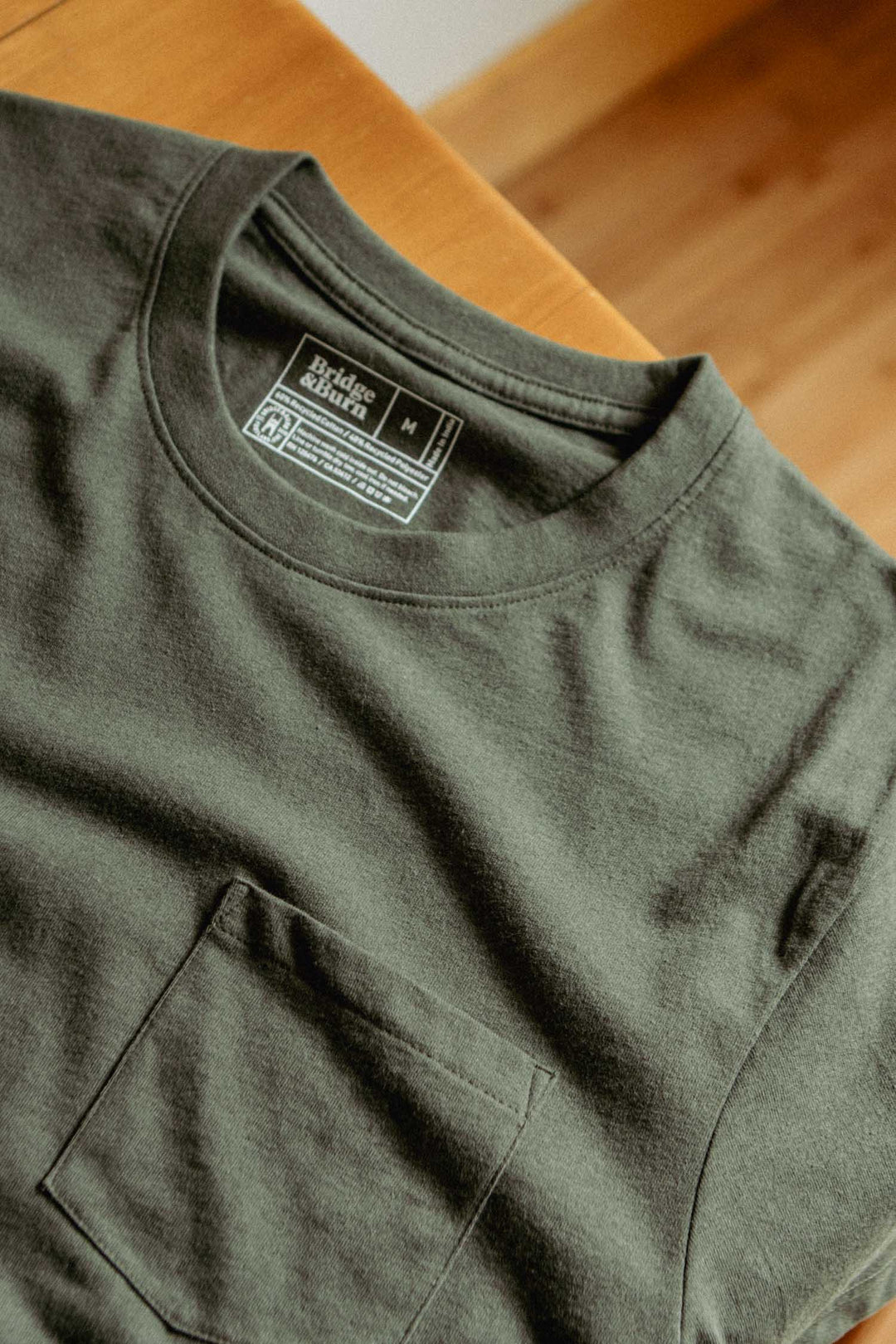 A close-up of the Bridge & Burn Recycled Pocket Tee in olive, featuring a chest pocket and visible collar tag, displayed flat on a wooden surface.