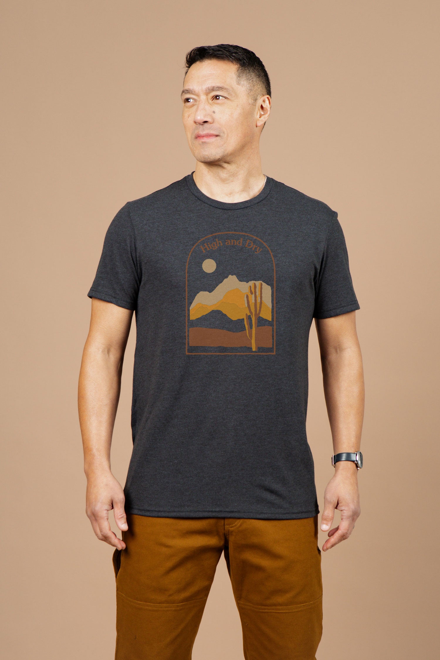 A man stands against a beige background wearing Bridge & Burn's Men's High & Dry Tee in black, featuring a desert mountain graphic and text, paired with brown pants. He looks slightly left and smiles softly.