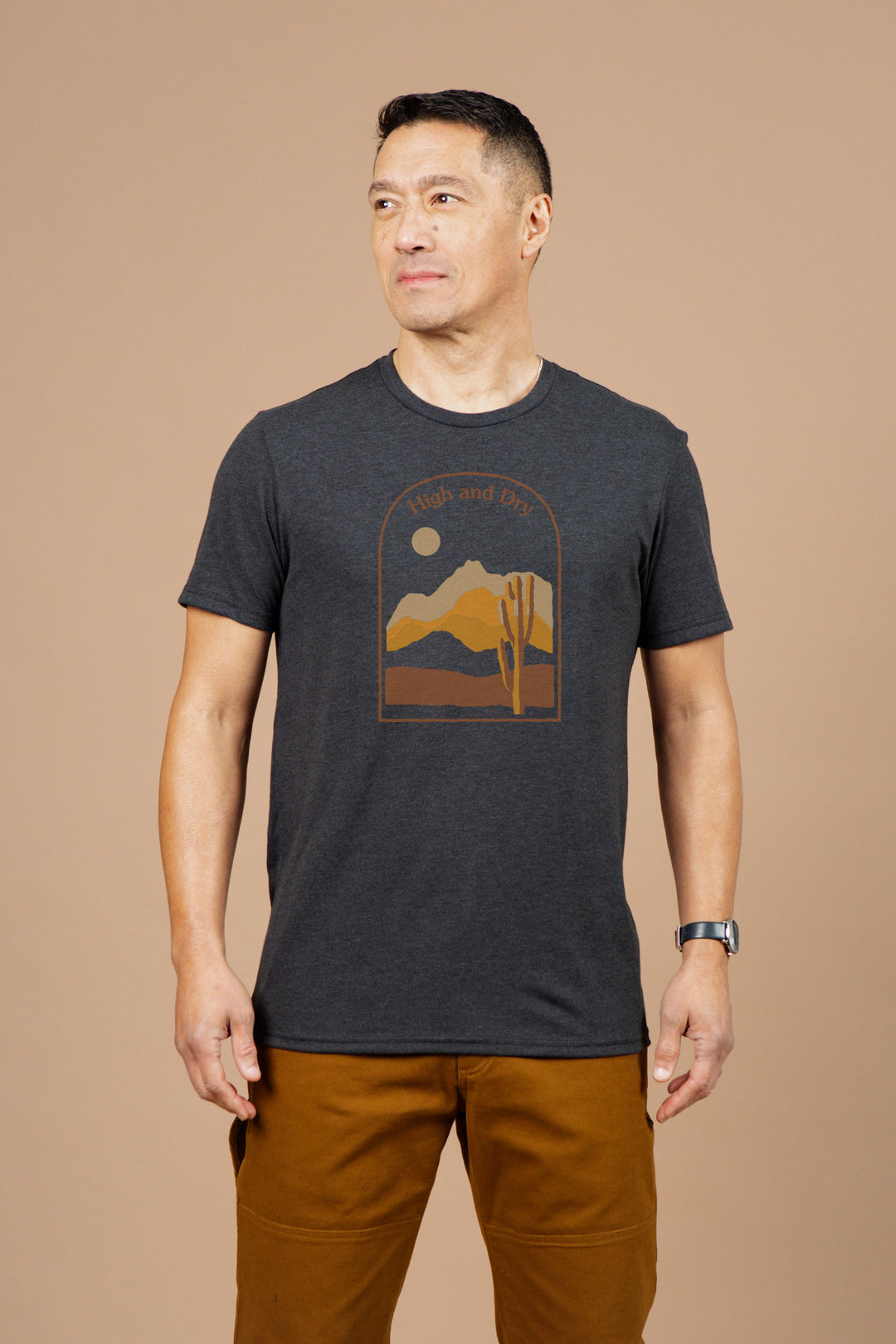 A man stands against a beige background wearing Bridge & Burn's Men's High & Dry Tee in black, featuring a desert mountain graphic and text, paired with brown pants. He looks slightly left and smiles softly.