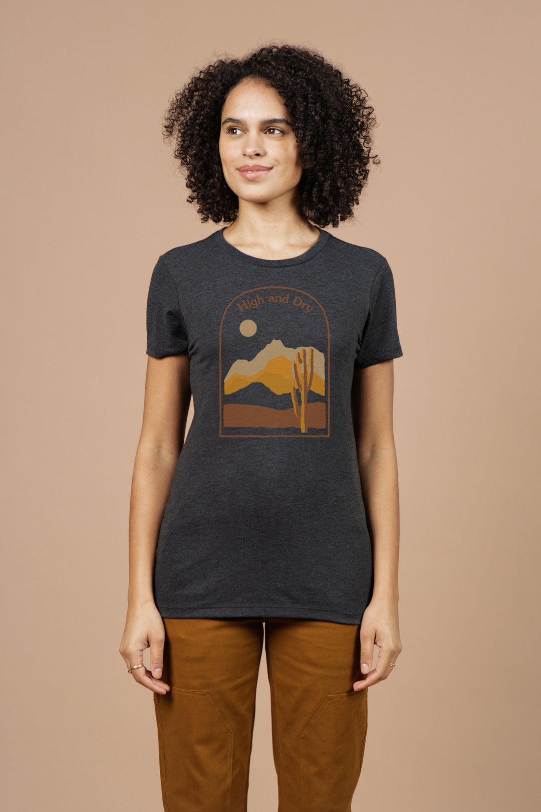 A woman with curly hair wears the Bridge & Burn Women's High & Dry Tee in black—an organic cotton shirt with a desert landscape and "High and Dry" text. She pairs it with brown pants against a plain light brown background.