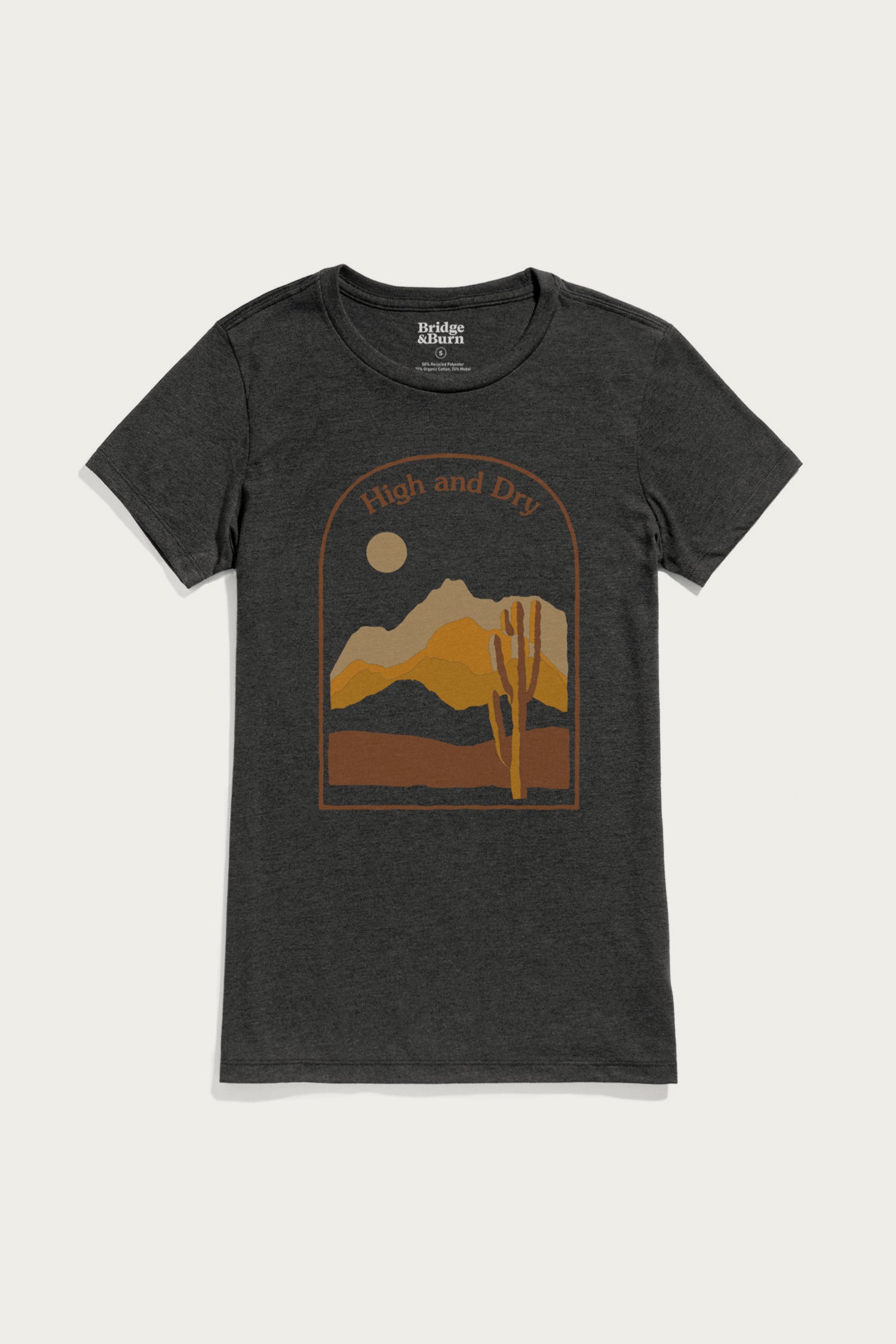 The Women's High & Dry Tee by Bridge & Burn is a black organic cotton shirt with an arched frame desert graphic—mountains, cactus, sun or moon—and "High and Dry" in yellow-orange tones.