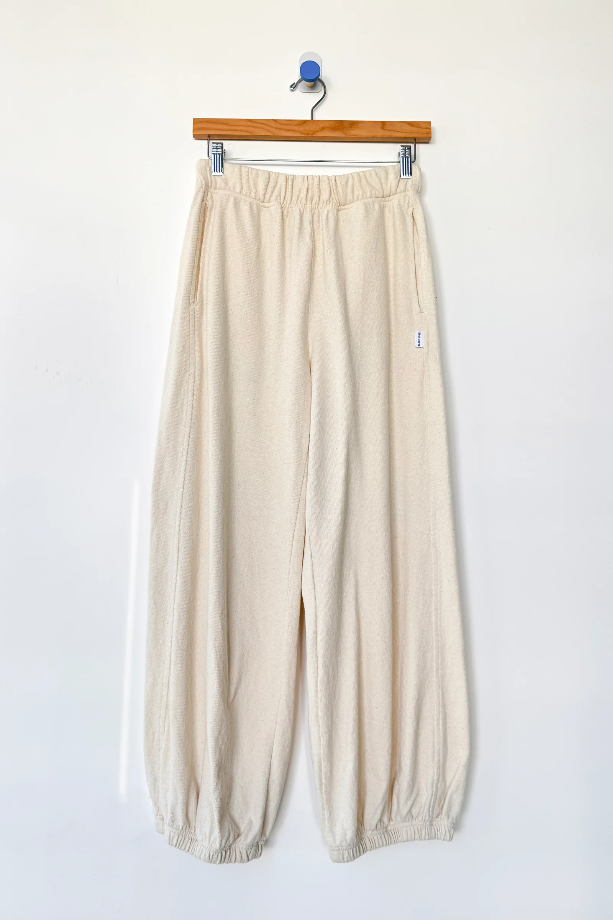 Le Bon Shoppe - Balloon Pant / Natural: Cream-colored, loose-fitting balloon leg pants made from undyed cotton with elastic waistband and cuffs by Le Bon Shoppe, shown hanging on a wooden hanger against a plain white wall.