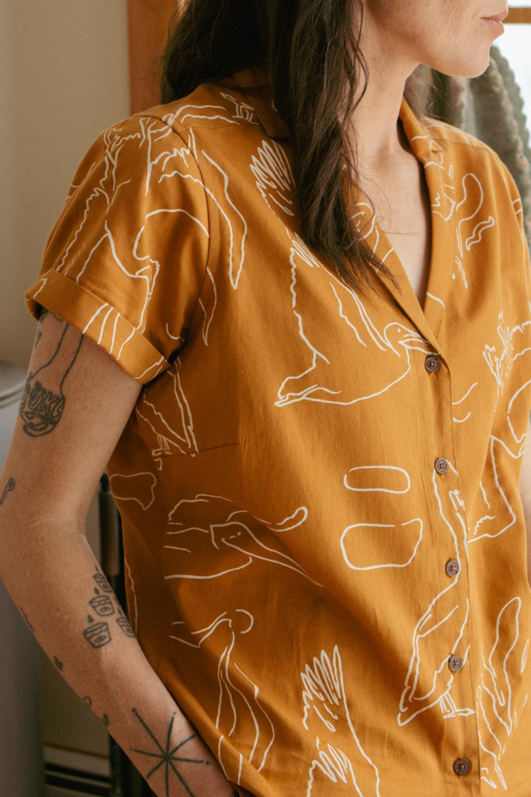 Person wearing the Bridge & Burn Innes Shirt / Shoreflight—a mustard, relaxed-fit button-up with abstract white bird designs. Their arm shows black tattoos and their dark hair is loose. Photo taken indoors in natural light.