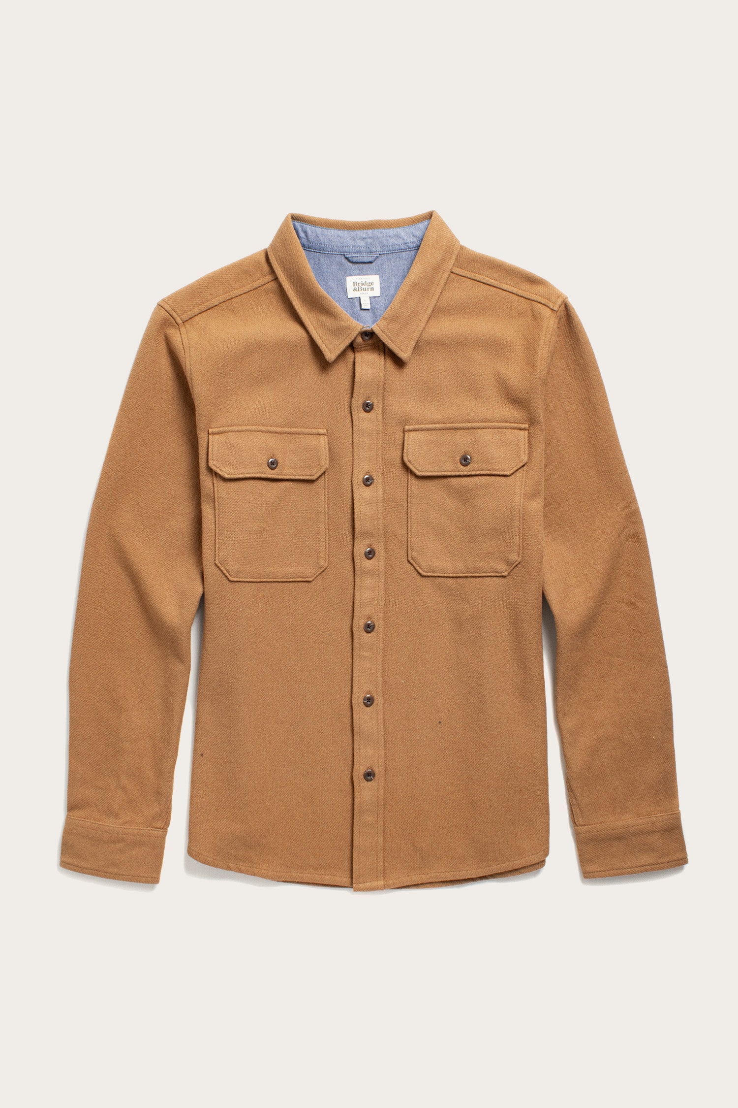 The Bridge & Burn Pointer Overshirt in camel is a tan twill style with long sleeves, two chest pockets, brown buttons, and a light blue fabric lining inside the collar—an eco-friendly and stylish choice. Displayed flat on a light background.