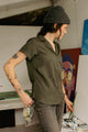Wearing the Bridge & Burn Innes Shirt in Olive and a gray beanie, a woman with arm tattoos stands in an art studio holding a paintbrush and cloth, surrounded by art supplies and a colorful painting.