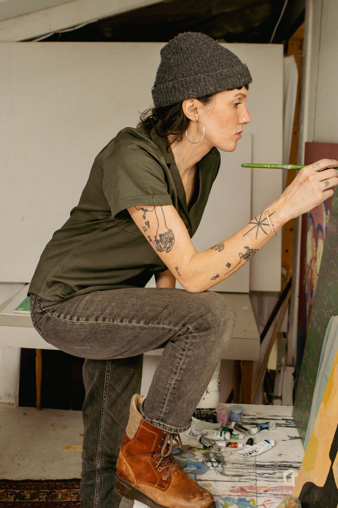 Kneeling in an art studio, a person with tattoos paints on canvas, wearing the Bridge & Burn Innes Shirt in Olive, a beanie, and hoop earrings—showcasing creative style and flair.