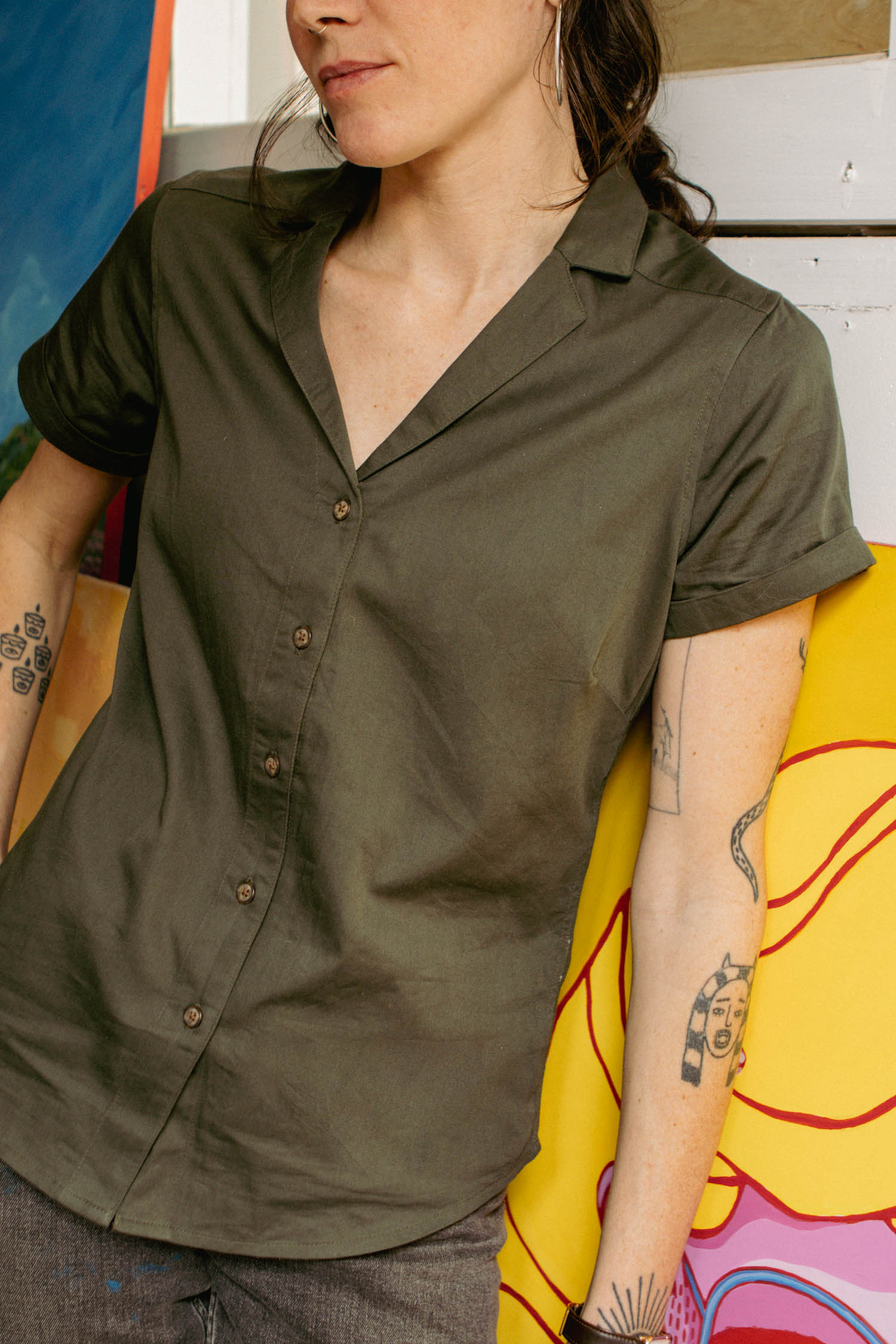 Wearing the Bridge & Burn Innes Shirt in Olive, a person stands beside a vibrant painted canvas. Their tattooed arm rests casually on the artwork, complementing their creative style.