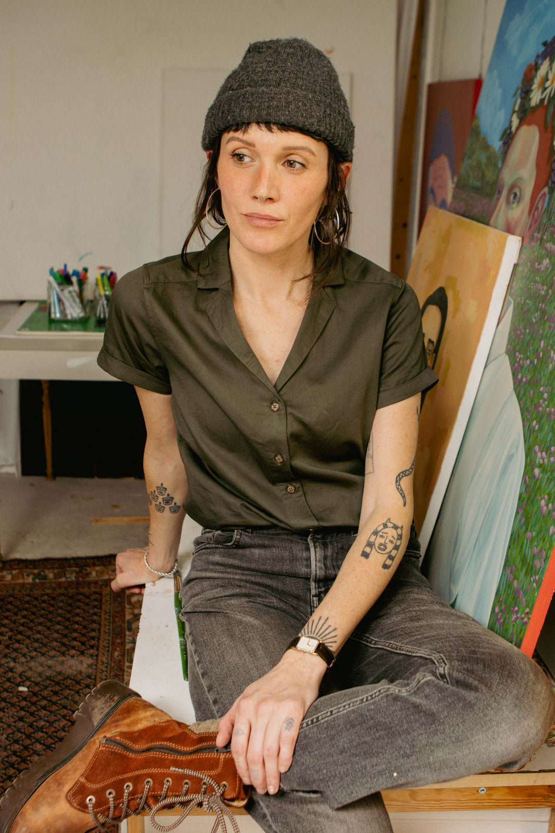 A woman in an olive Innes Shirt by Bridge & Burn, a gray beanie, and gray jeans sits on a table beside colorful portrait paintings. She has tattoos on her arms and gazes thoughtfully away from the camera.
