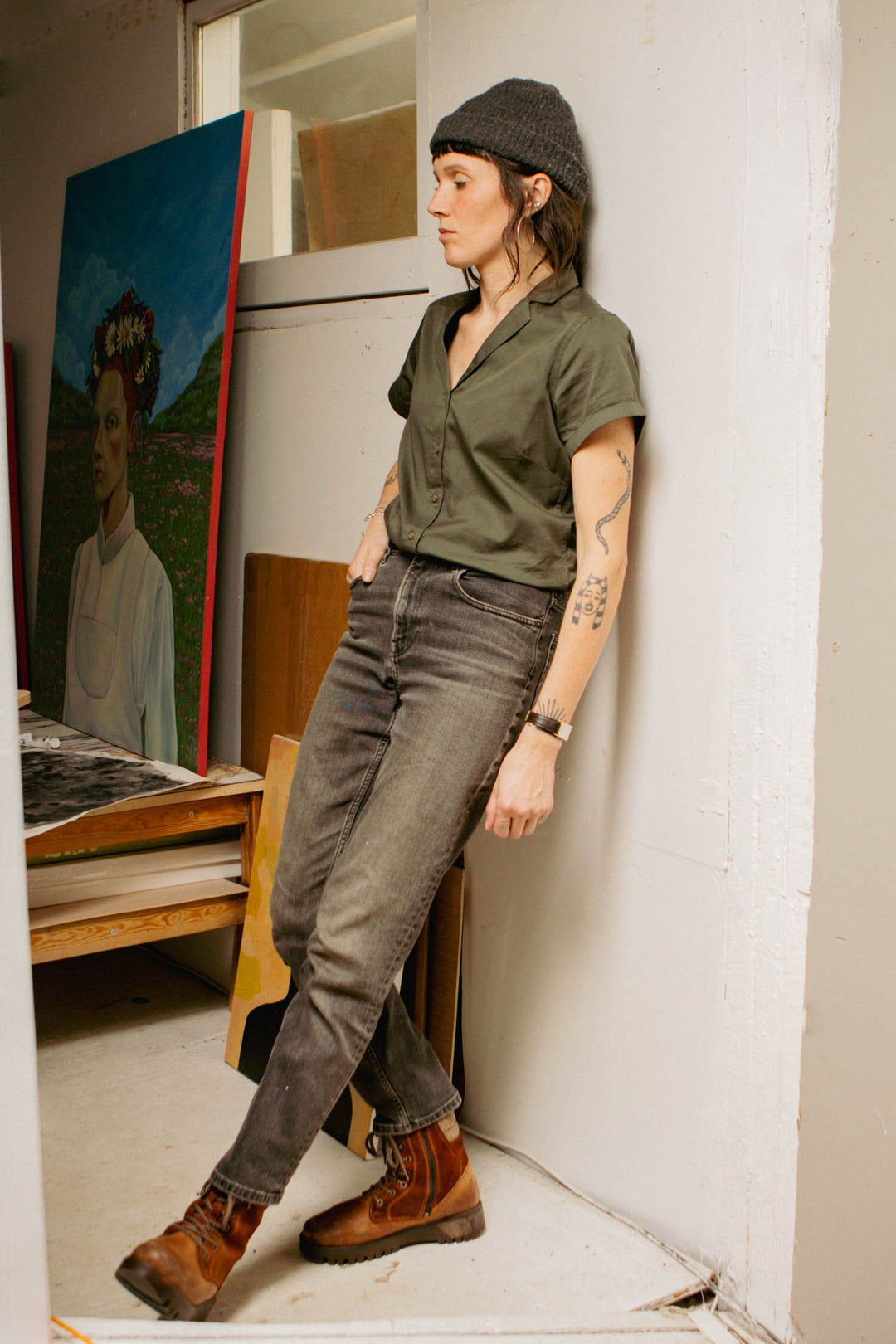 A tattooed person in a gray beanie, Bridge & Burn Innes Shirt in olive, faded jeans, and brown boots leans against a wall in an art studio with paintings, including a portrait of a woman wearing a flower crown, behind them.