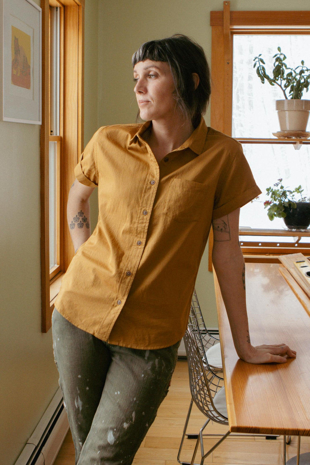 A person with short dark hair and tattoos leans on a wooden table by a window, wearing the Bridge & Burn Bea Shirt in Gold and olive pants. Sunlight pours in while houseplants rest on shelves near the window.