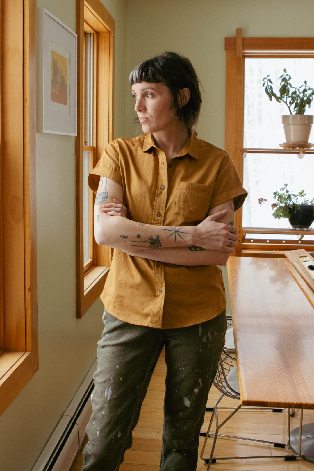 A person with short dark hair and tattoos stands indoors by a window, arms crossed, wearing the Bridge & Burn Bea Shirt in Gold and green paint-stained pants. Wooden accents and plants are visible in the background.