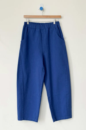 The Le Bon Shoppe - Arc Pant in Azure is a loose-fitting, high-waisted pant with an elastic waistband and side pockets, shown on a wooden hanger against a plain white wall.