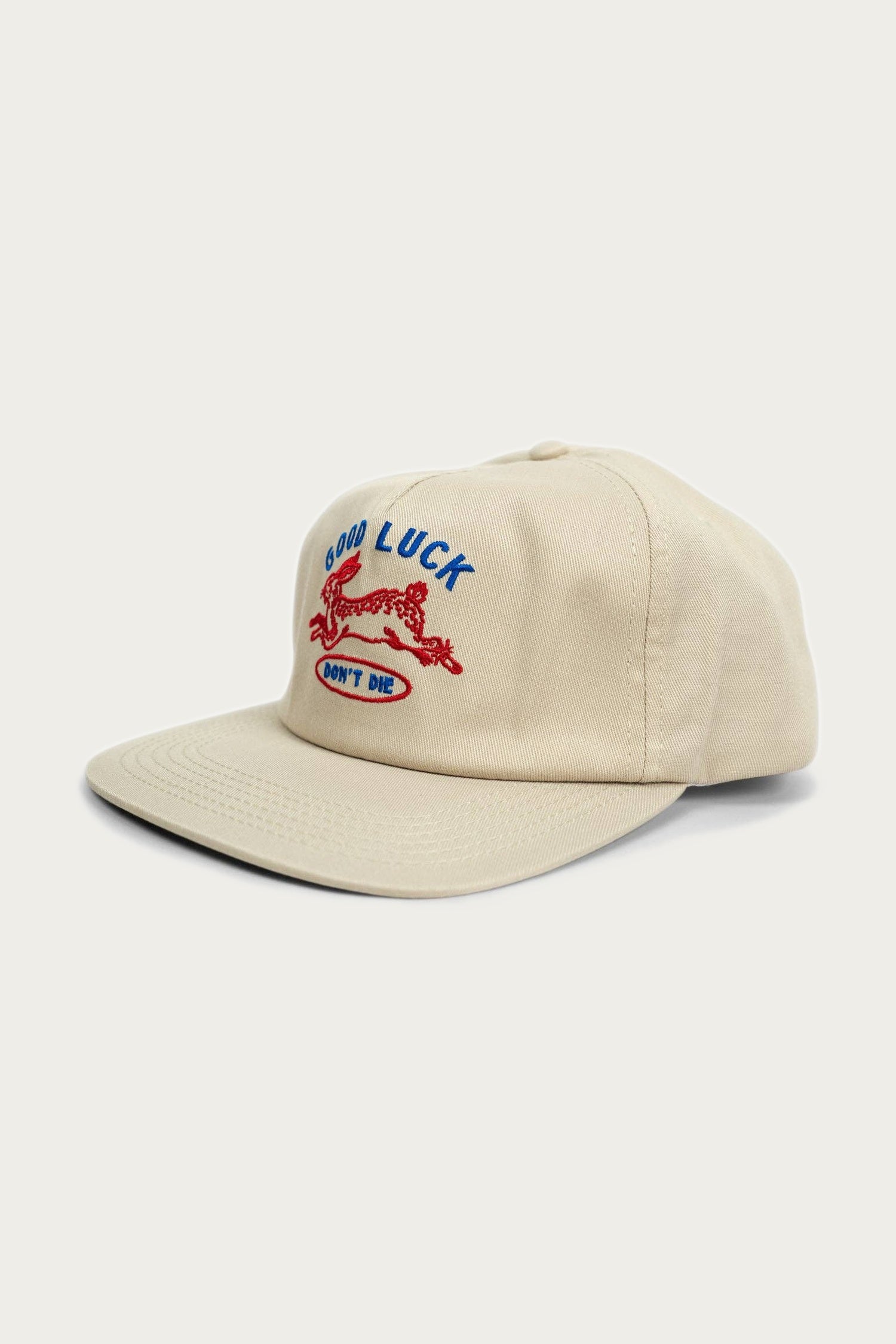 The Iron & Resin Good Luck Hat in tan features front embroidery with a red running dog, blue GOOD LUCK text, and a white DONT DIE patch. This cotton twill snapback hat from Iron & Resin is adjustable for a comfortable fit.