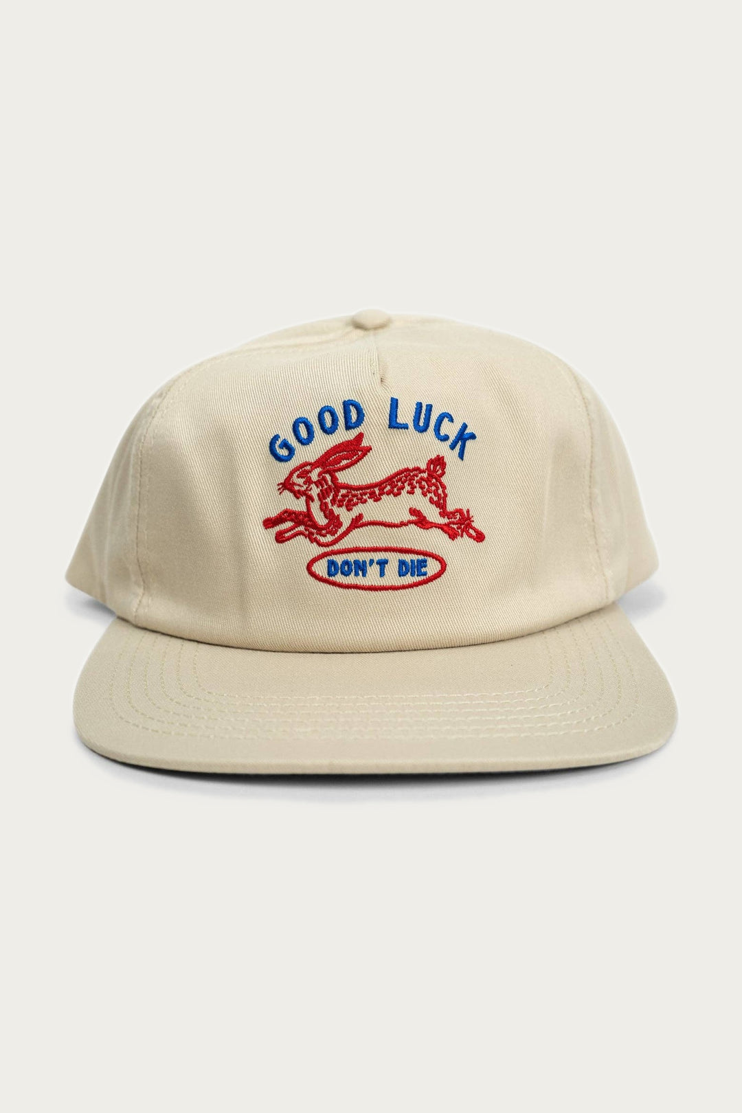 The Iron & Resin - Good Luck Hat / Tan is a beige cotton twill hat with a red embroidered running rabbit, blue "GOOD LUCK" text, and a red "DONT DIE" oval on the front. It features an adjustable snapback for the perfect fit.