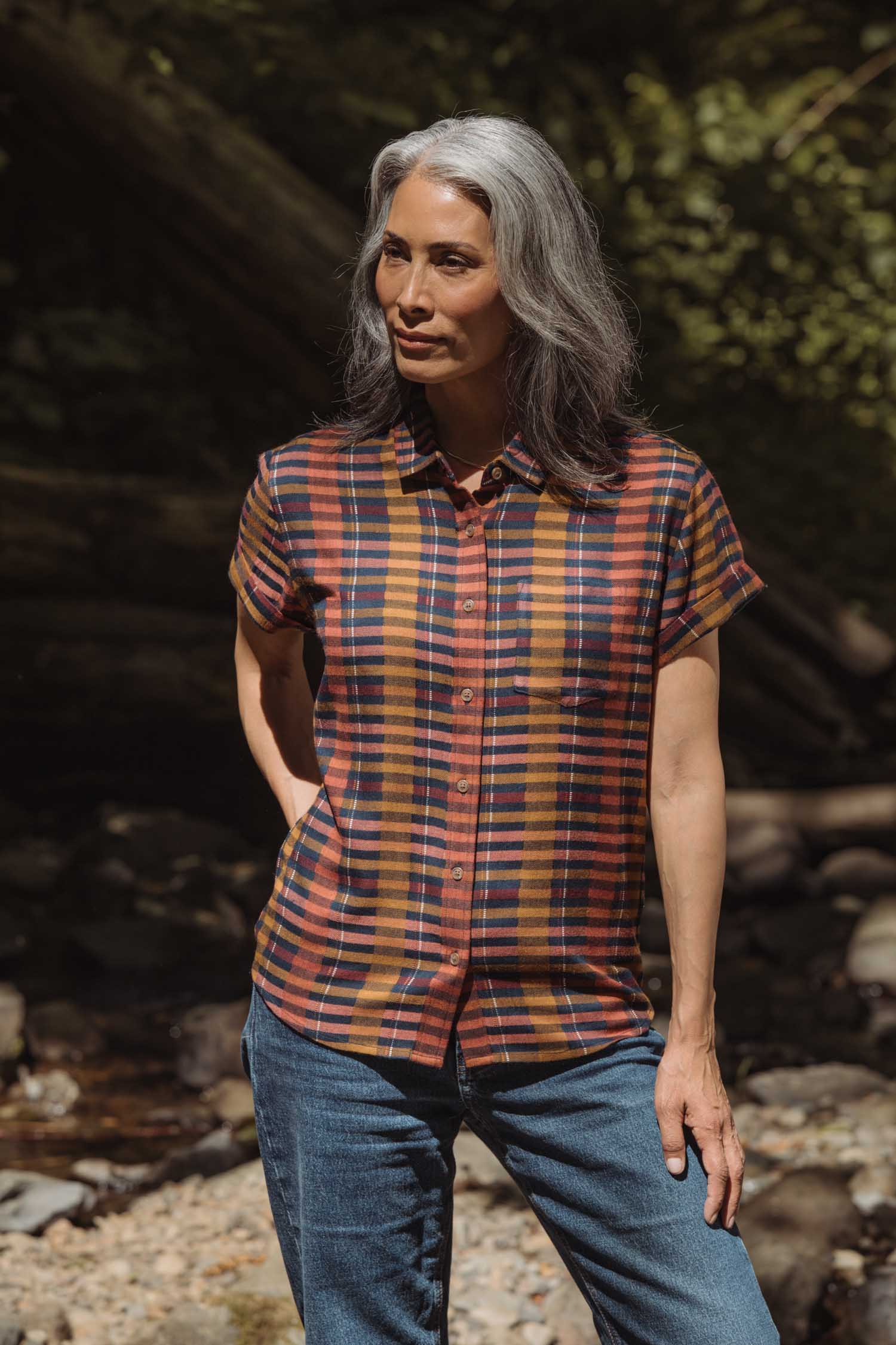 A woman with long gray hair stands outdoors in sunlight, wearing the Bridge & Burn Bea Shirt in Alpenglow Stripe and blue jeans. She looks to the side with one hand in her pocket.