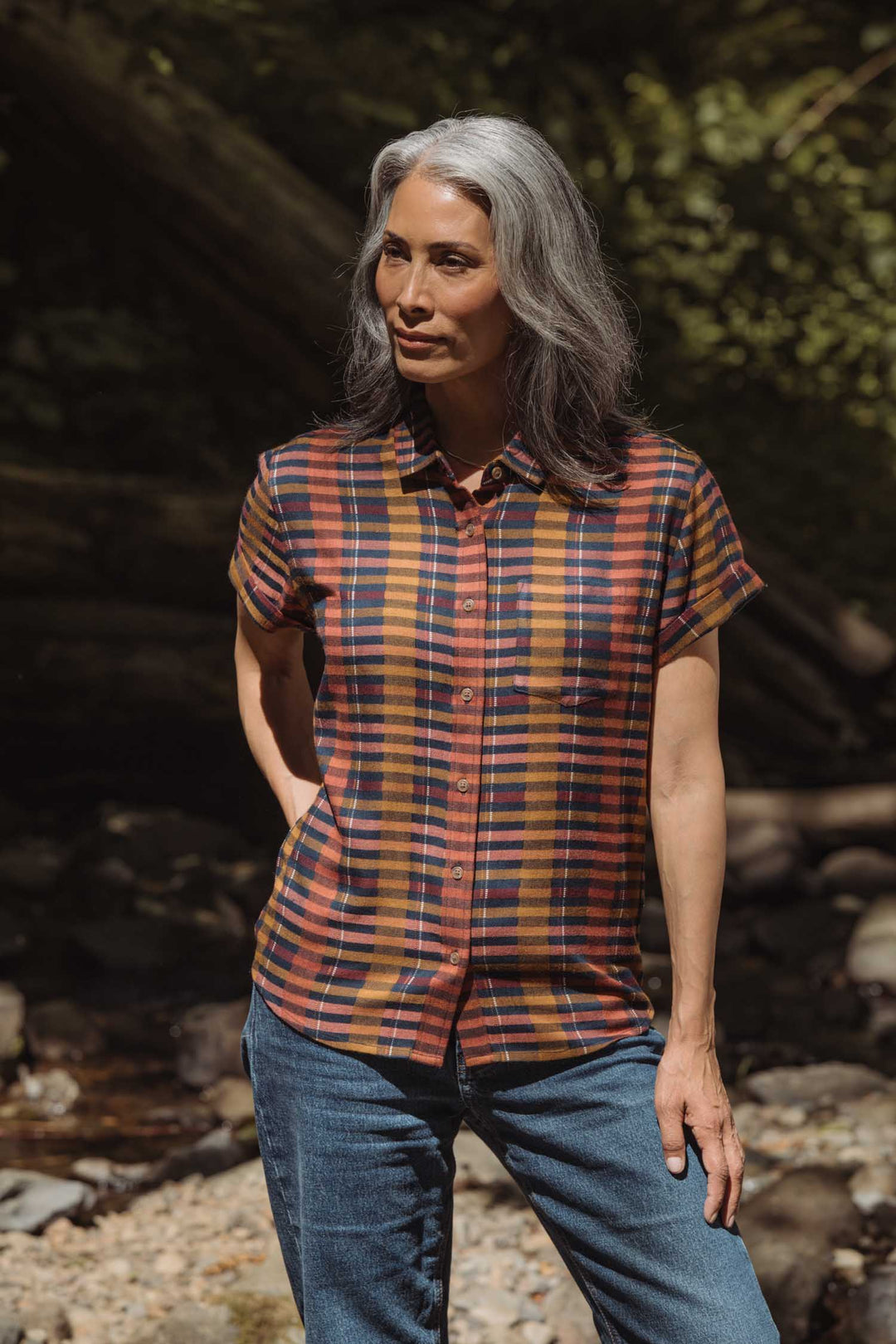 A woman with long gray hair stands outdoors in sunlight, wearing the Bridge & Burn Bea Shirt in Alpenglow Stripe and blue jeans. She looks to the side with one hand in her pocket.