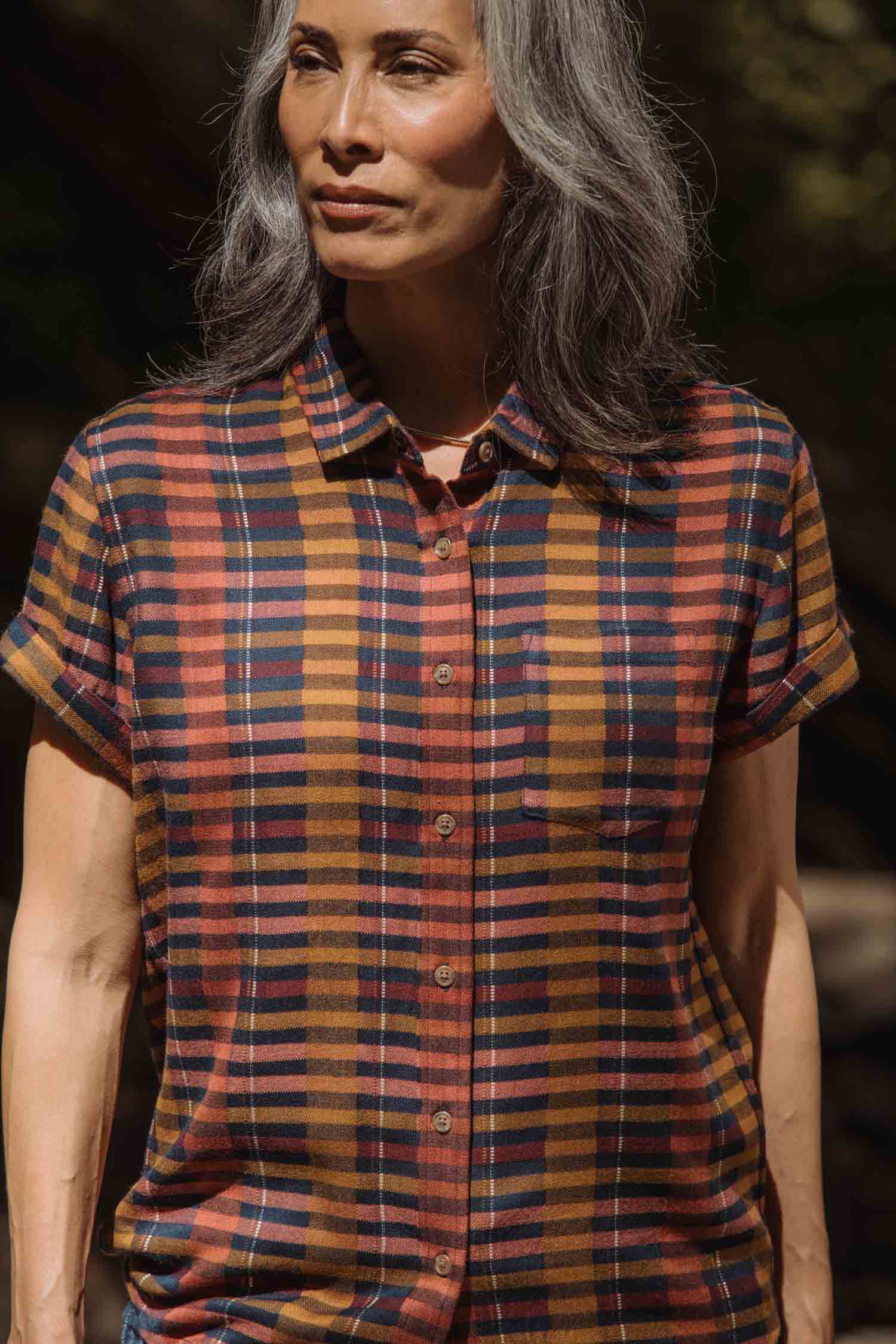 A person with long gray hair wears the Bridge & Burn Bea Shirt in Alpenglow Stripe, a short-sleeve button down made from an eco-friendly cotton alternative, standing outdoors in natural light.